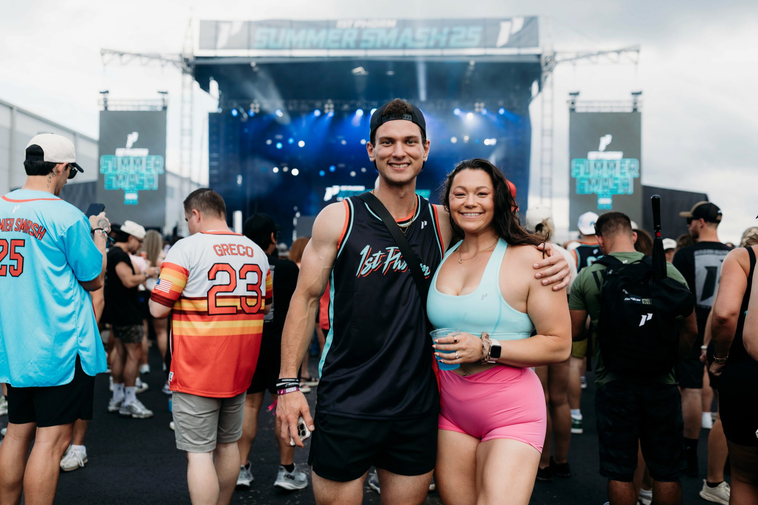Two smiling people, a man and a woman, standing together at an outdoor event with a stage in the background.
