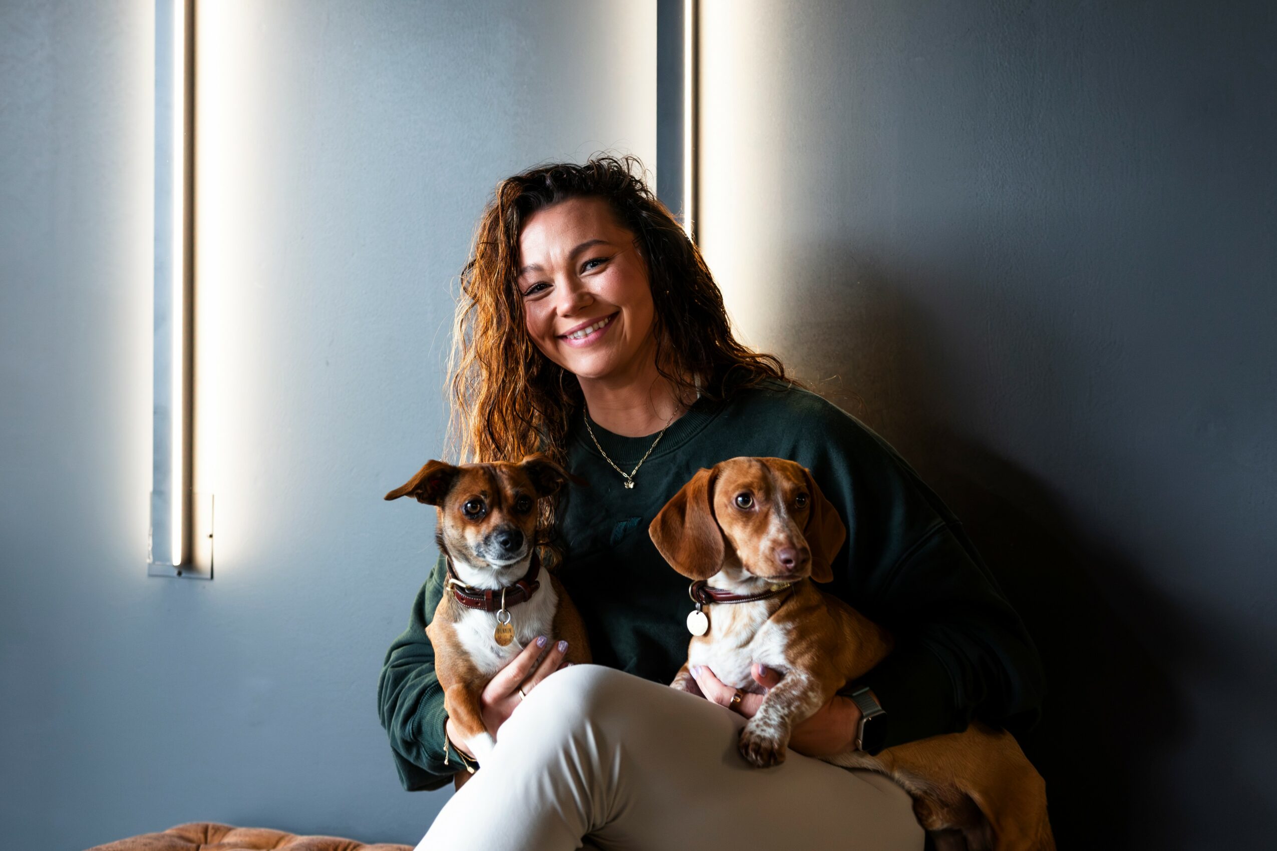 Woman with curly hair smiling, holding two small dogs, one in each arm, sitting indoors near a window with vertical lights.