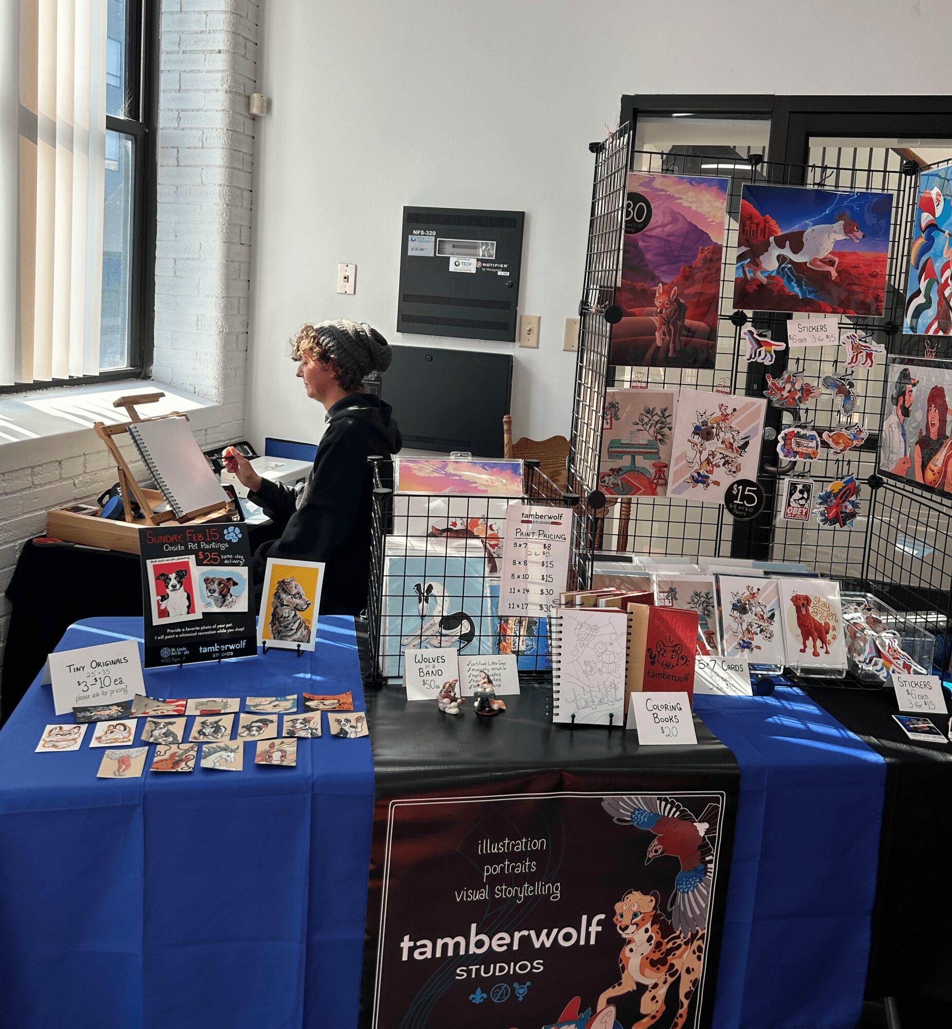 Child standing at an art display booth with artwork and cards on tables, and a wire grid with colorful art pieces behind.