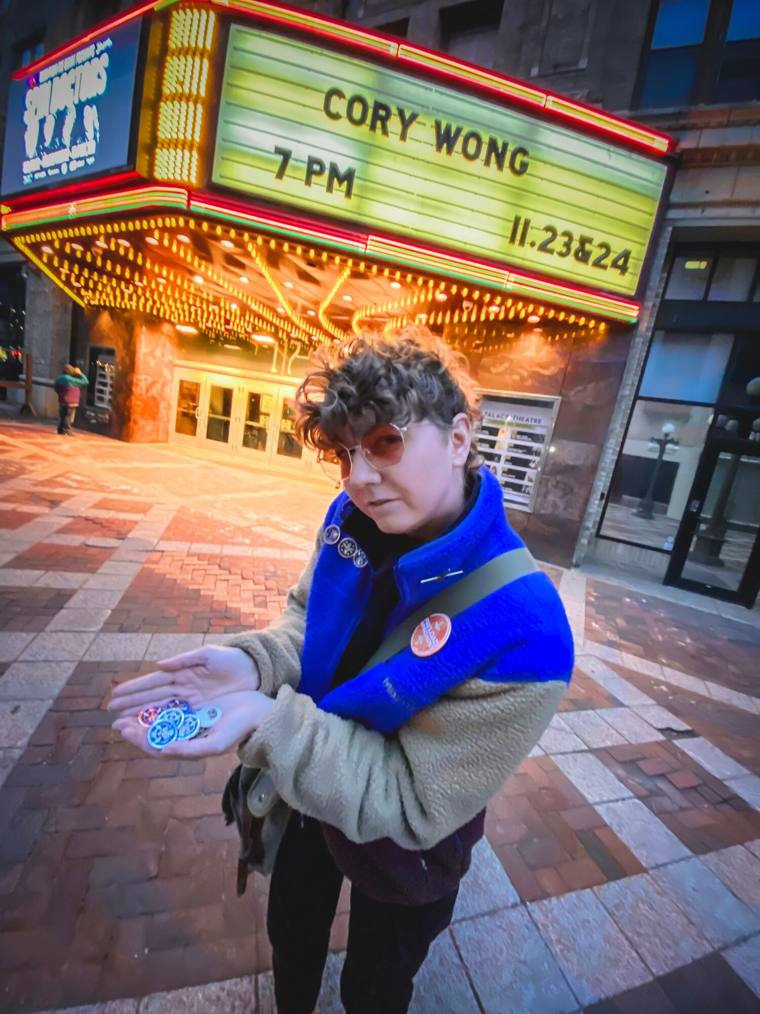 Young person with curly hair holding objects in hands, standing outside near a theater marquee displaying event details.