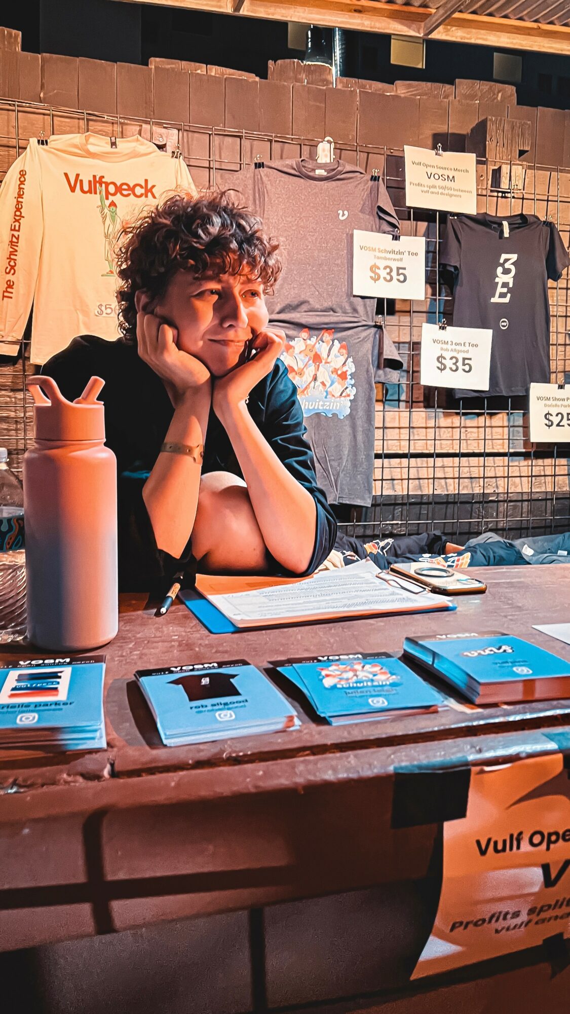 Young person with curly hair sitting at a table with papers and a water bottle, behind a display with clothing and signs.
