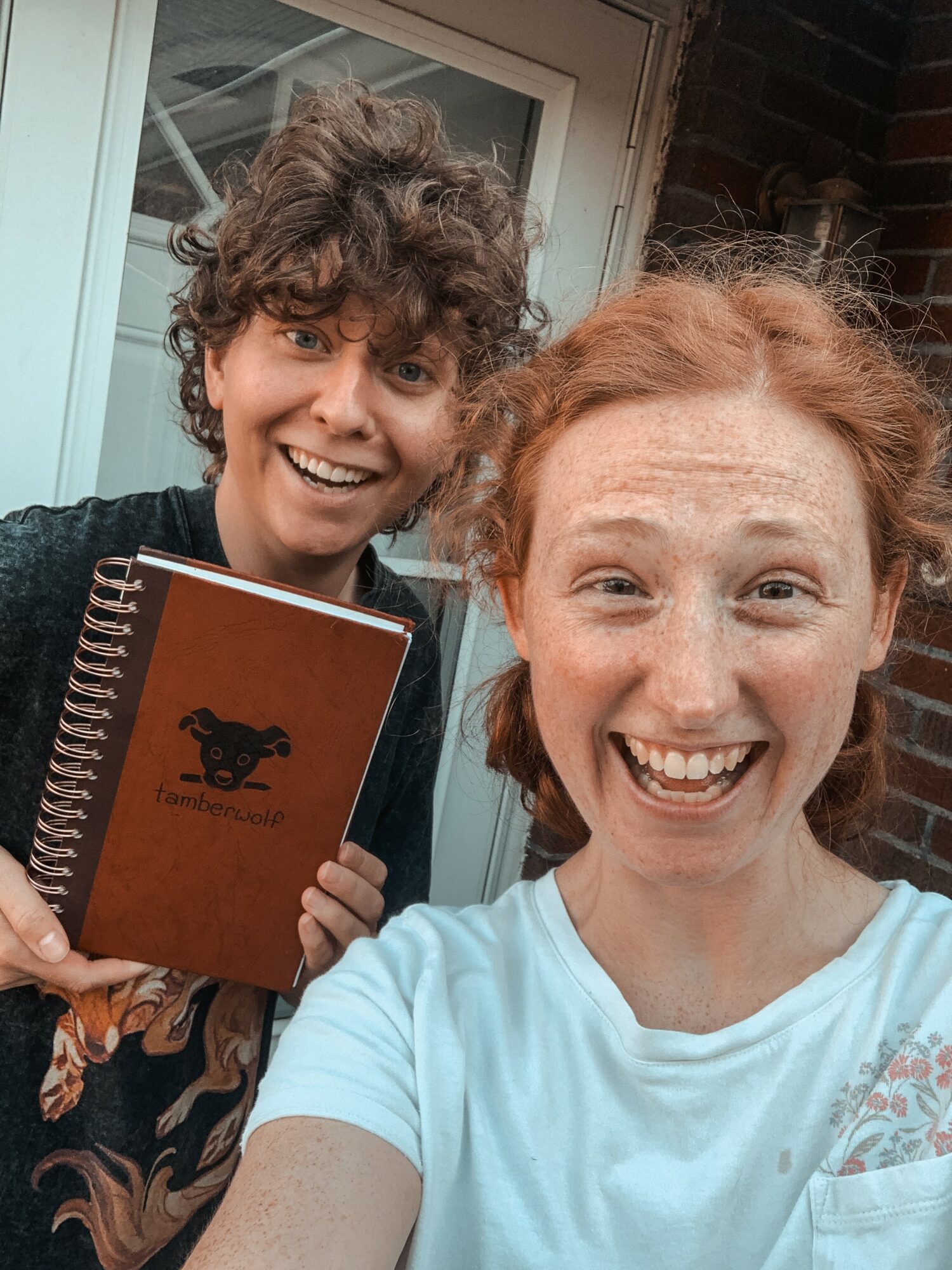 Two smiling women taking a selfie outside a building, one holding a notebook, both with curly hair and casual clothing.