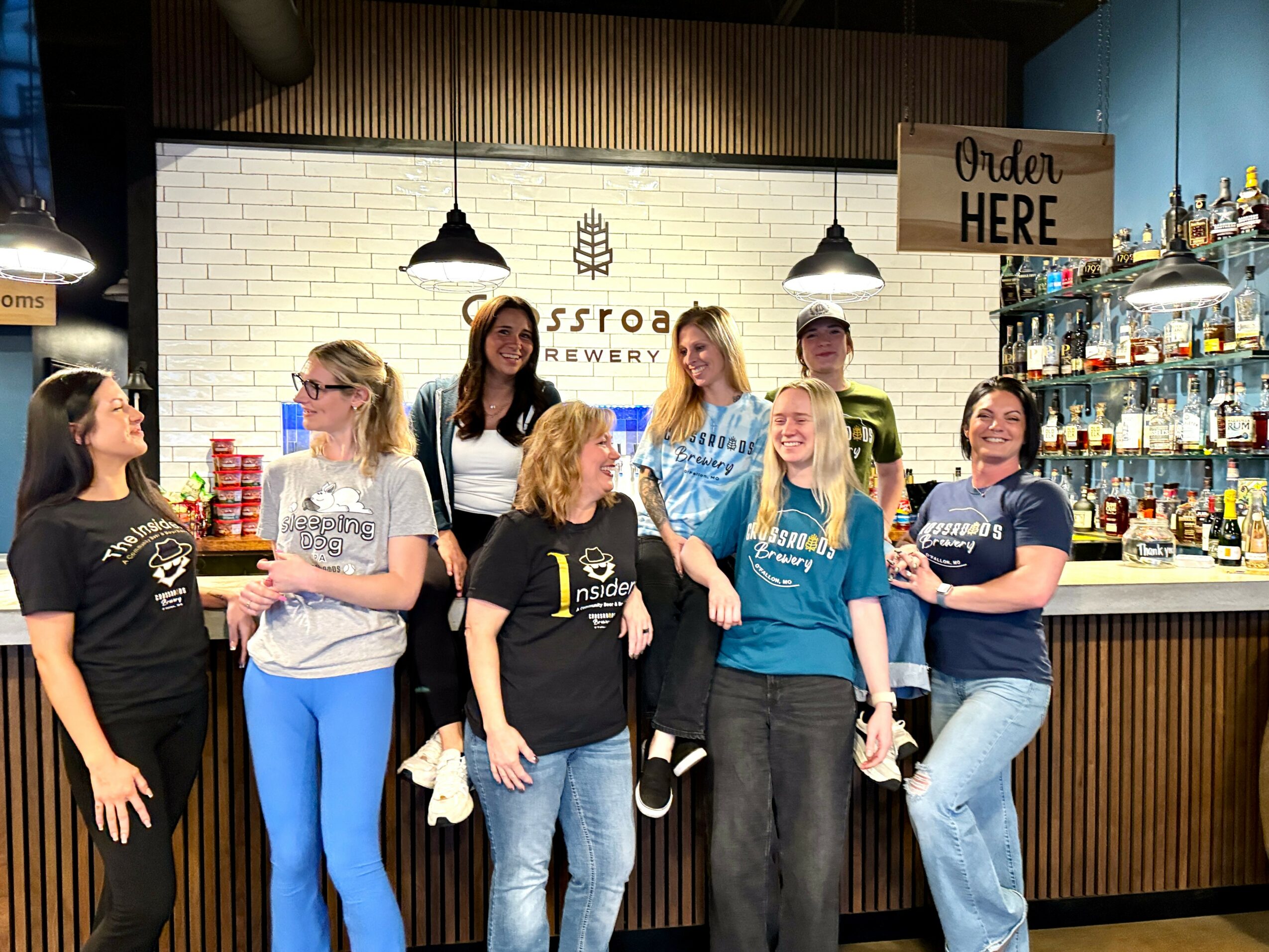 Group of seven women standing inside a brewery, smiling and talking, with a bar and shelves of bottles behind them.