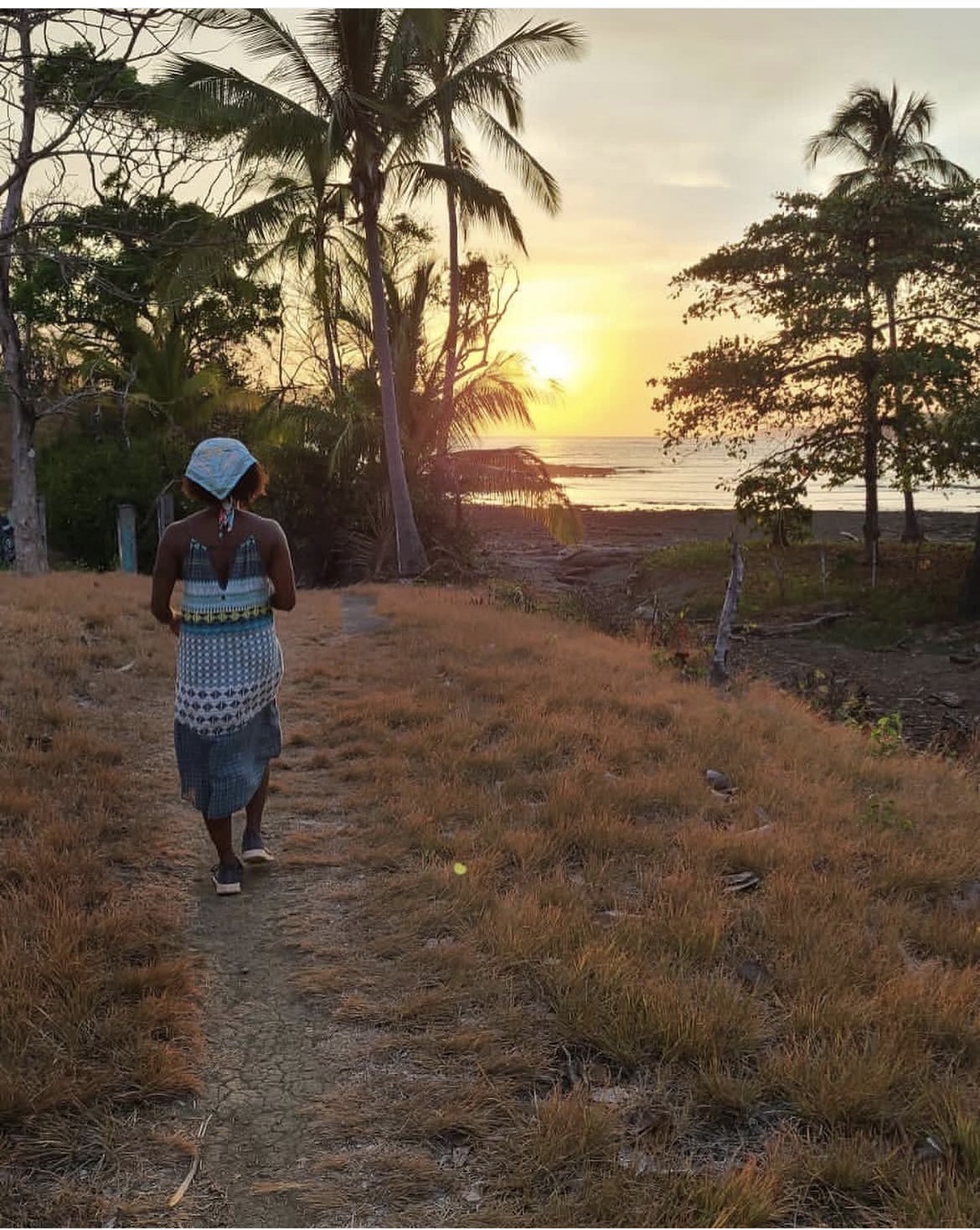 Woman walking along a dirt path near palm trees at sunset, with a beach in the background.