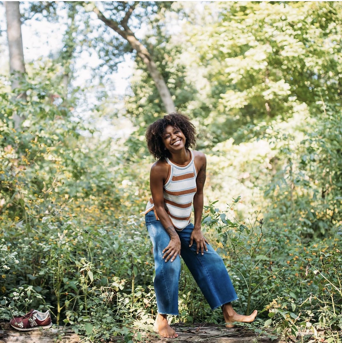 Young girl with curly hair smiling outdoors among trees and greenery, wearing a striped tank top and jeans, barefoot.