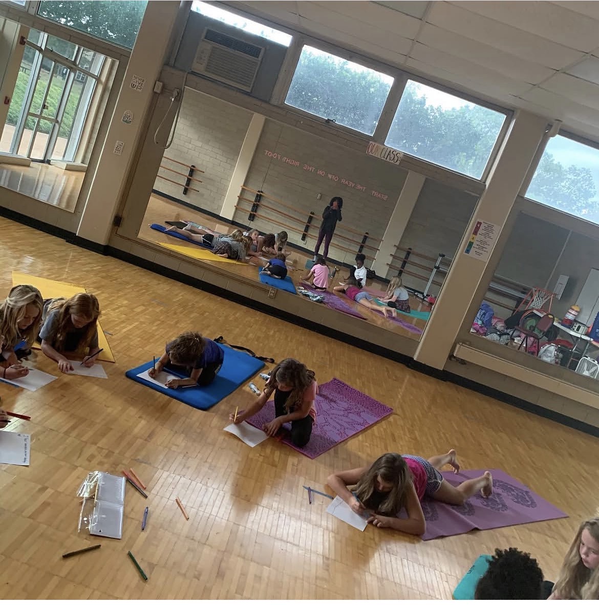 Children lying on mats writing or drawing in a classroom with large windows and a mirror reflecting the room.