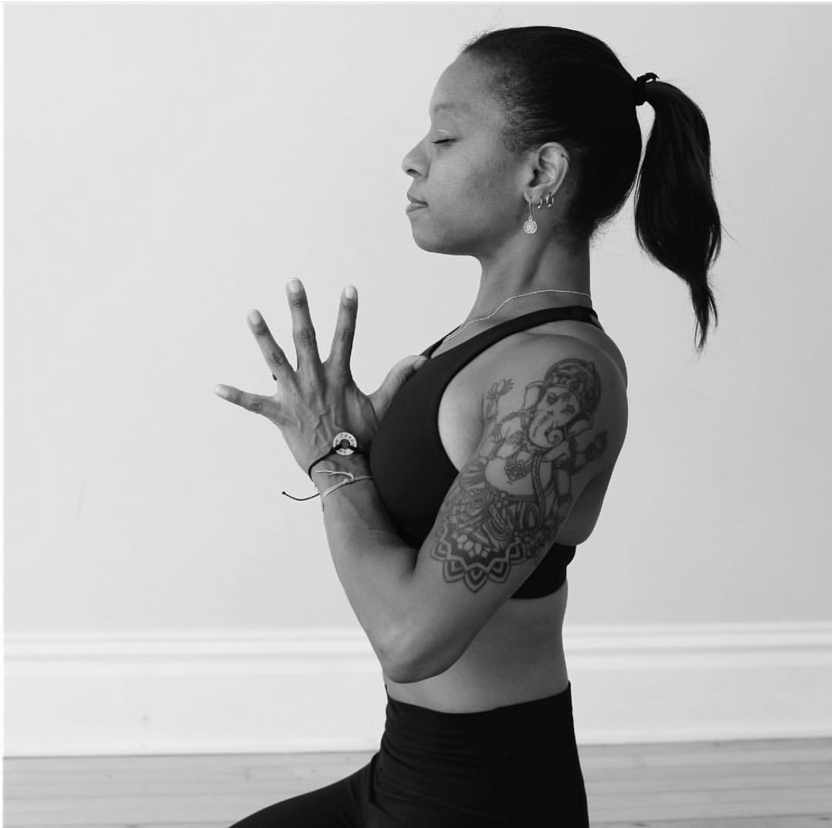 Woman practicing yoga with hands together, eyes closed, side profile, tattoos on arm, black sports bra, ponytail, plain background
