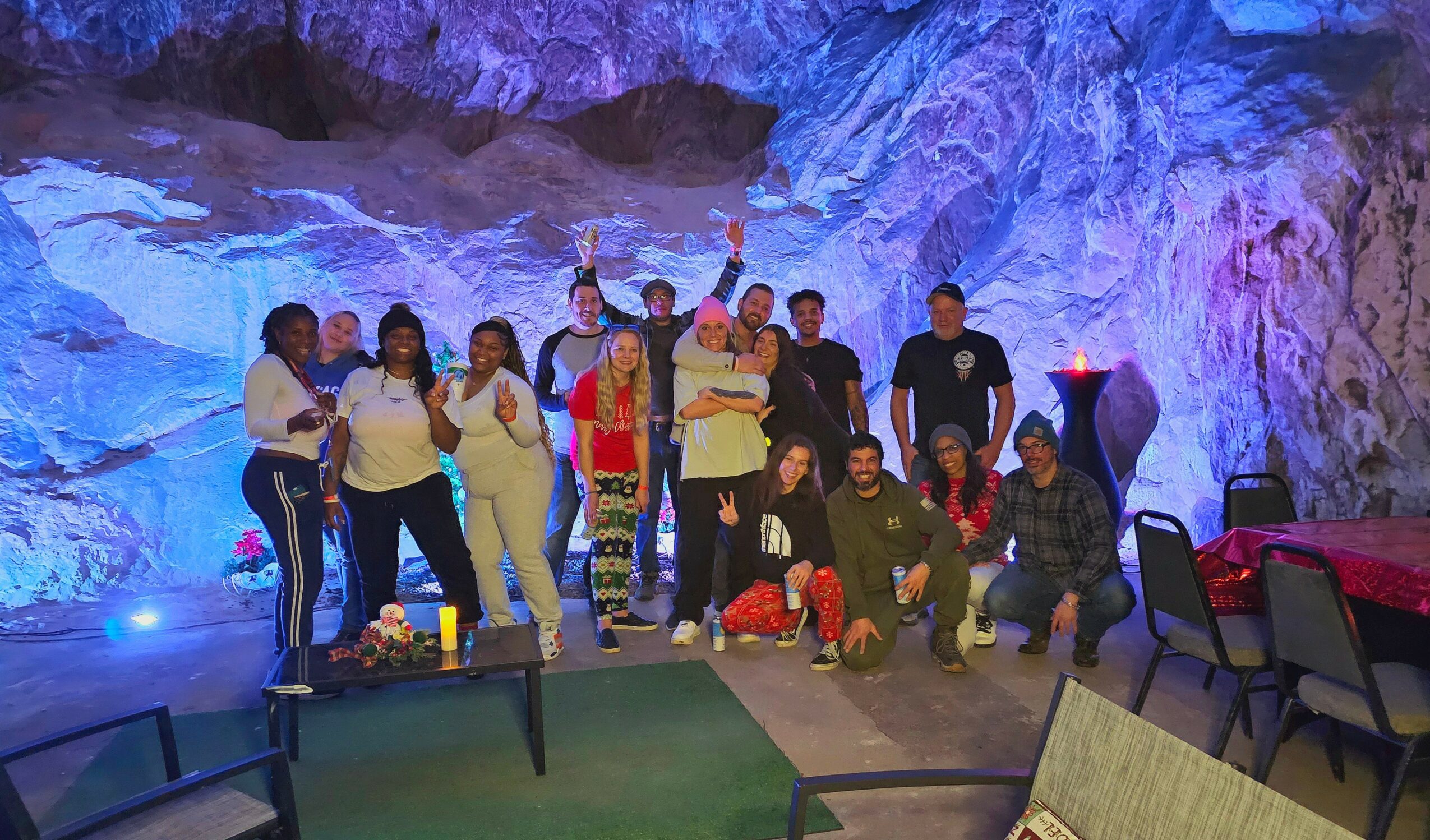Group of people posing in a cave with colorful lighting, some standing and some kneeling, with tables and chairs nearby.