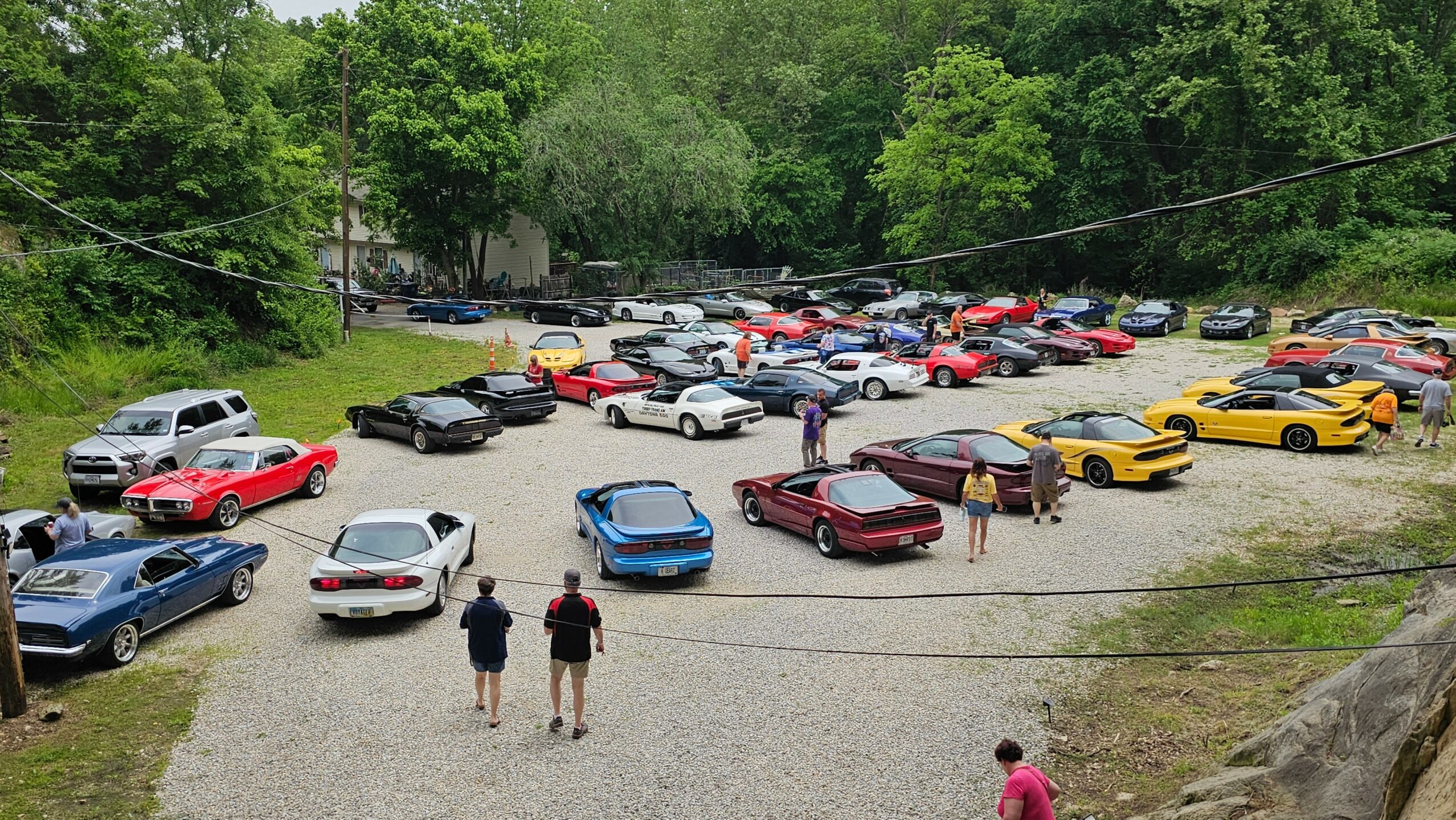 Parking lot filled with various cars, trees in background, two people walking towards the lot, one person standing nearby.
