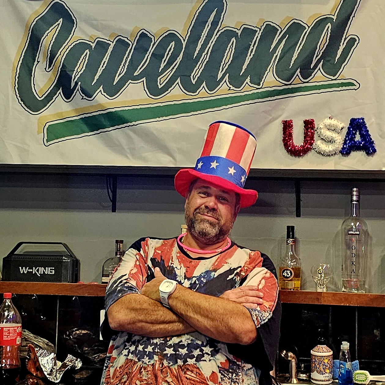 Man with beard wearing a patriotic hat and shirt, crossing arms, standing in front of a bar with bottles and glasses, USA sign, Cleveland banner.