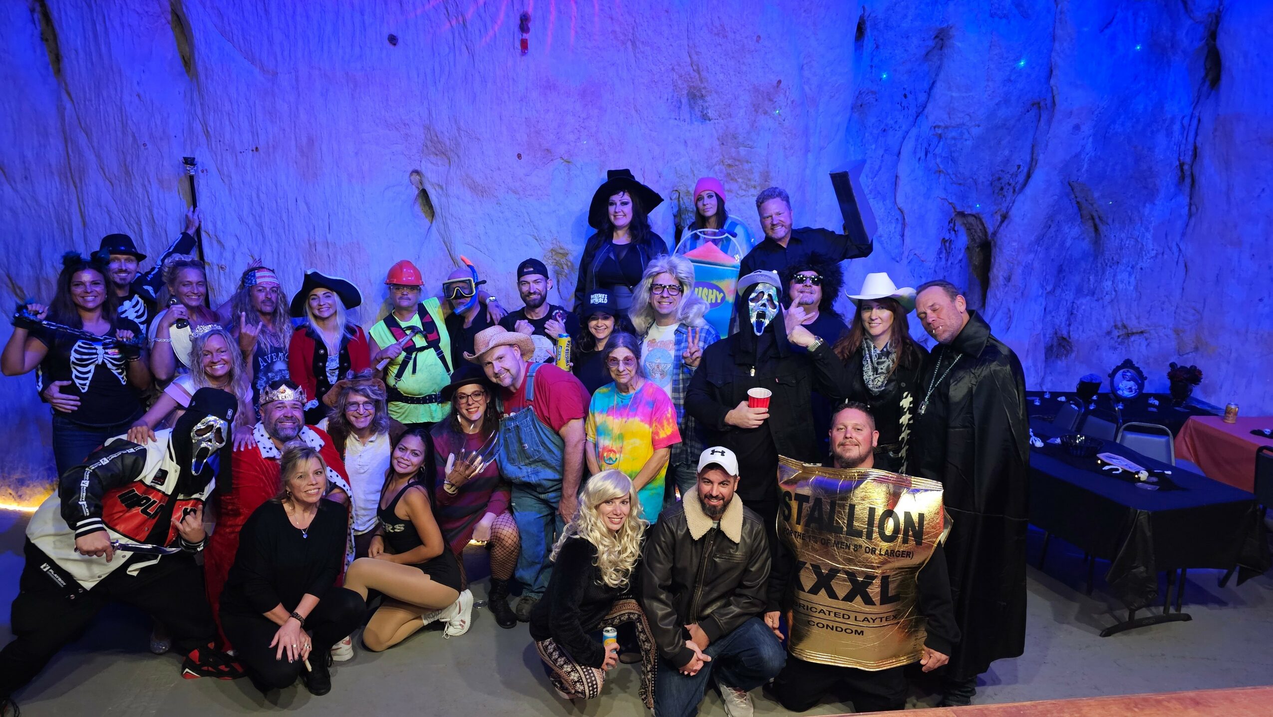 Group of people in costumes and masks posing indoors with a rocky background, some holding props or signs.