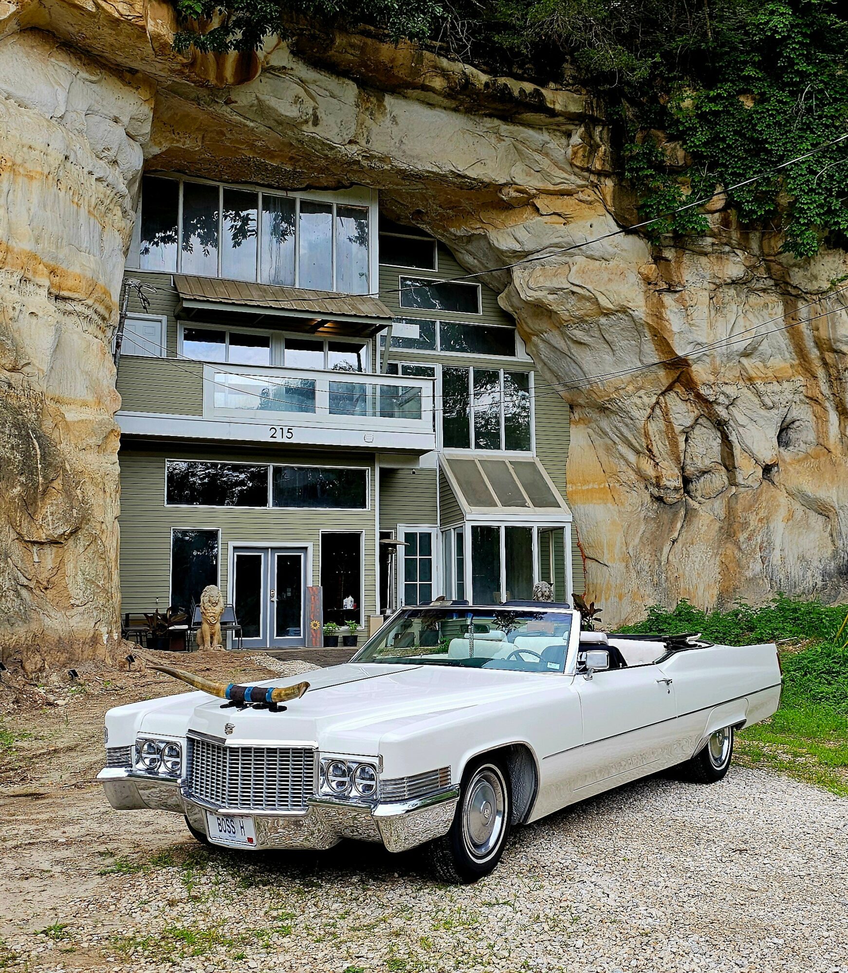 White vintage convertible car parked in front of a modern house built into a rocky cliffside, with greenery around.