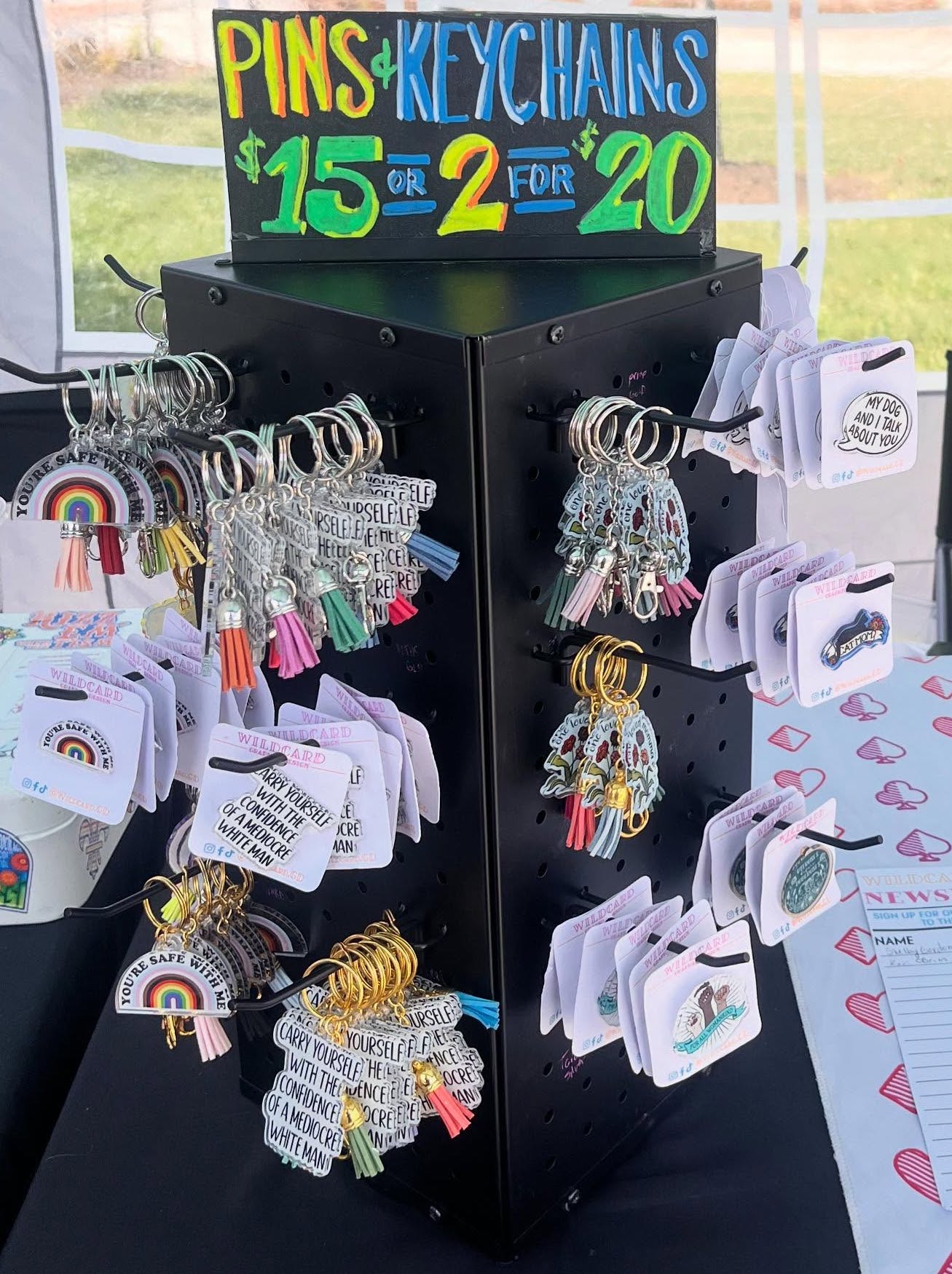 Display stand with colorful pins and keychains, price signs, and accessories on a table outdoors.