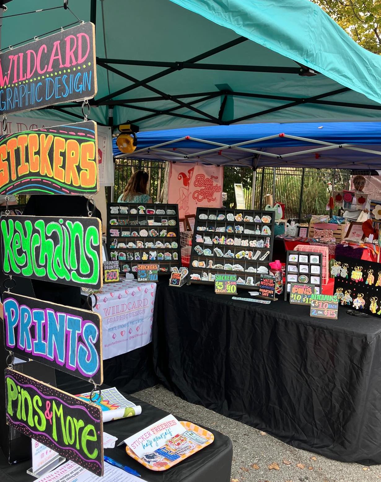 Outdoor market stall with signs for wildcards, stickers, keychains, prints, pins, and more, under a green canopy.