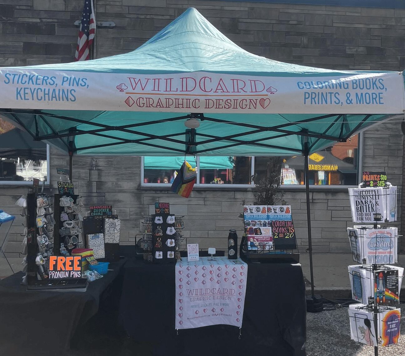 Outdoor booth with a blue canopy selling stickers, pins, keychains, and books, with a stone wall background.