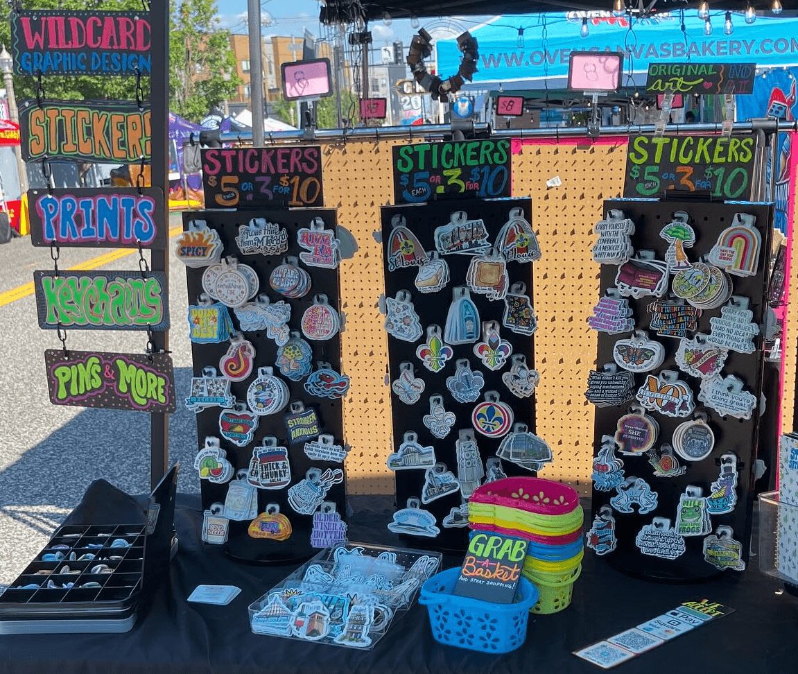 Display of colorful stickers, pins, and prints at an outdoor market stall with signs and a pegboard backdrop.