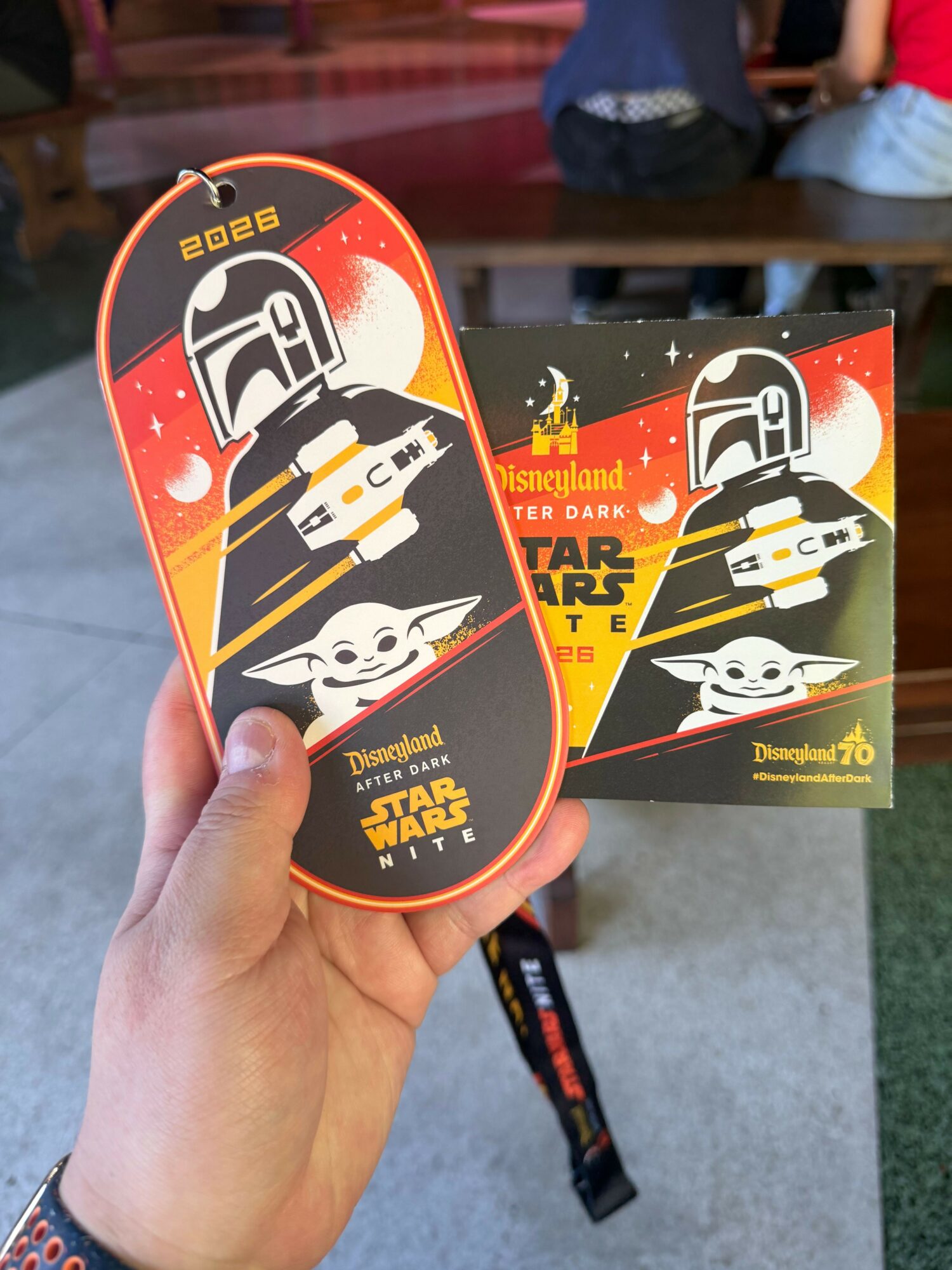 Hand holding a Star Wars-themed pin and card featuring a cartoon Mandalorian and Baby Yoda, with a Disneyland logo, in a park setting.