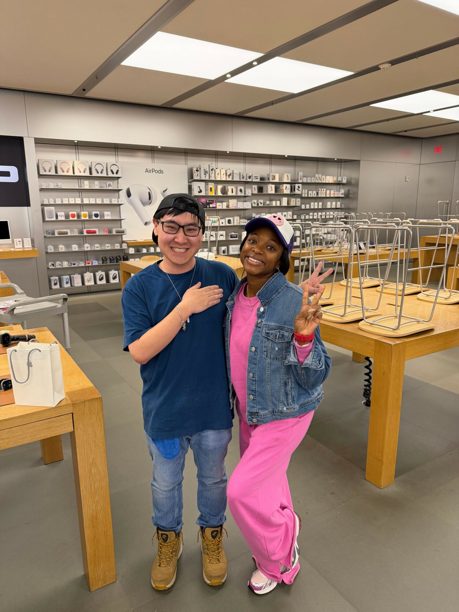 Two people standing inside a store, smiling and making peace signs, with shelves of electronic accessories in the background.