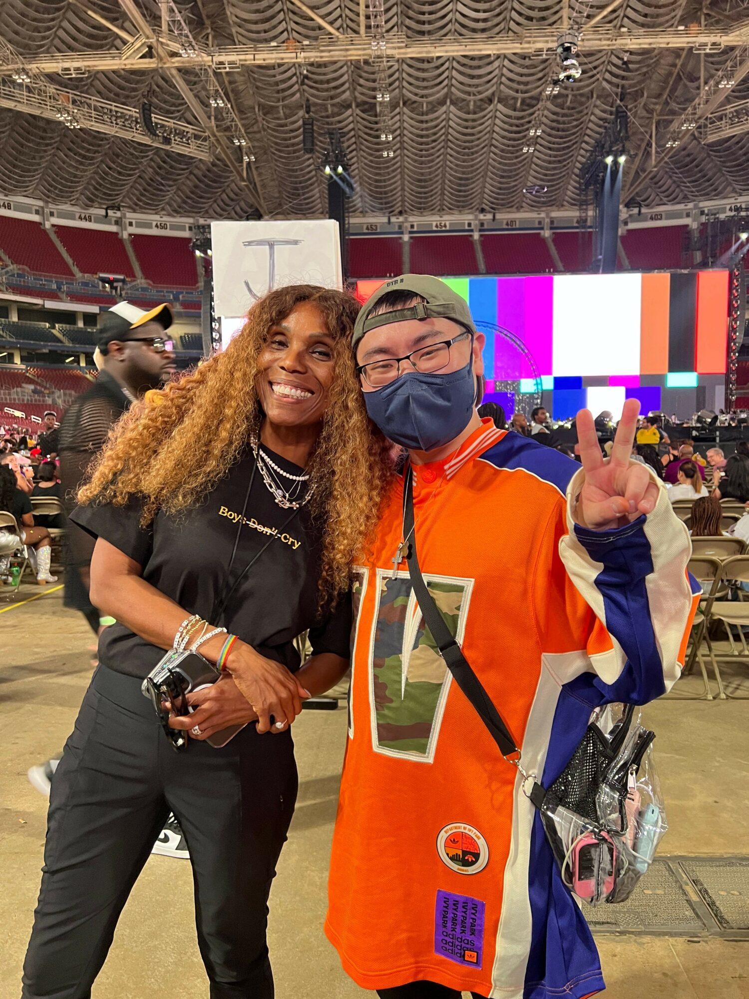 Two people smiling and posing for a photo at an indoor event, one wearing a black outfit and the other in an orange sports jersey with a face mask.
