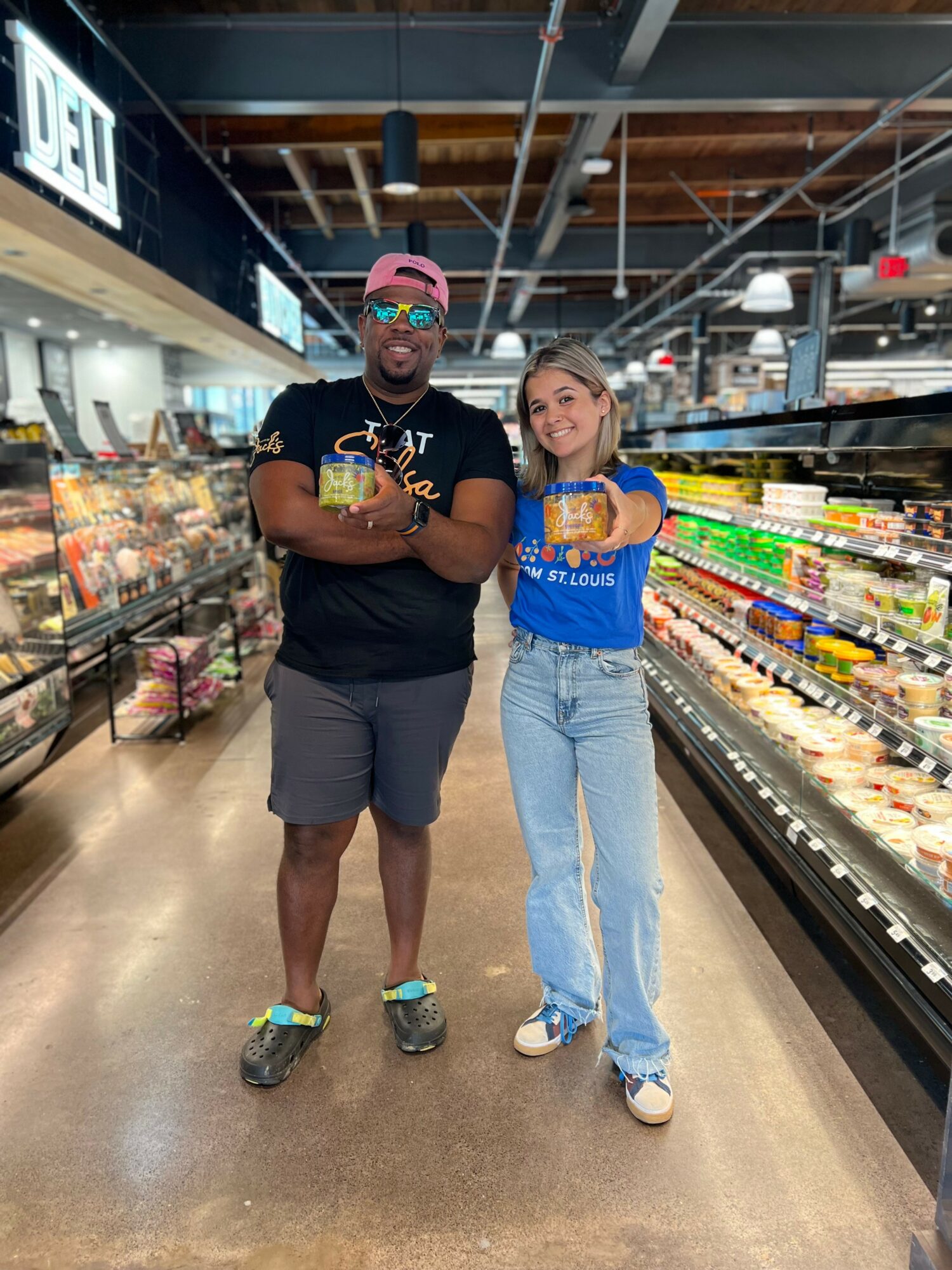 A man and a woman stand together in a grocery store aisle, smiling at the camera, holding drinks.