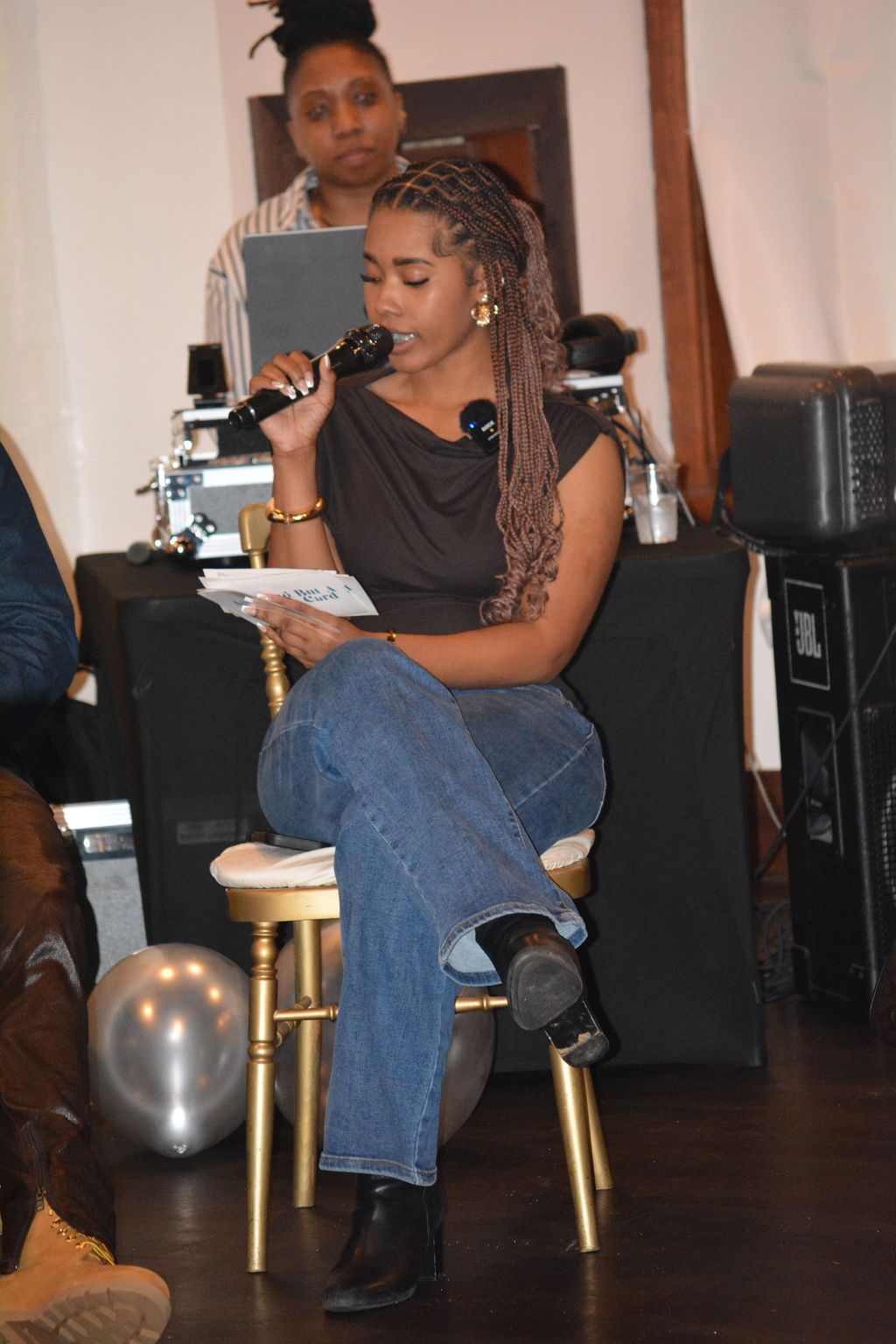 Woman sitting on a chair, holding a microphone and reading from a paper, with a woman standing behind her in the background.
