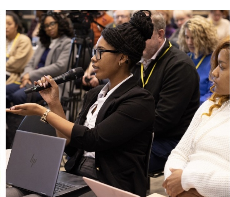 Woman speaking into microphone at a conference, with laptop in front, surrounded by seated audience members.