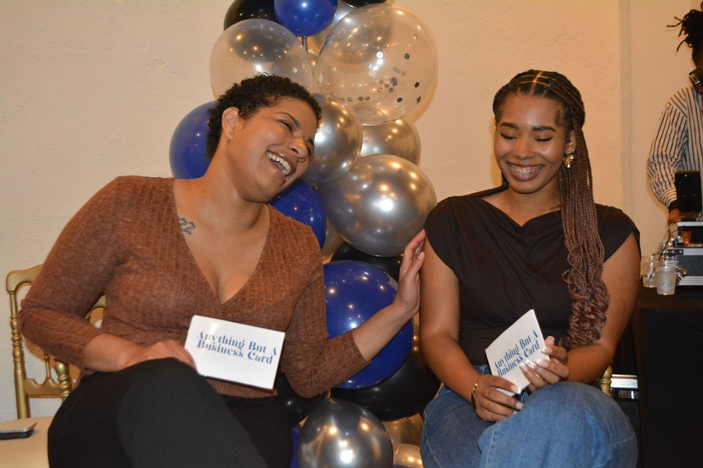 Two women smiling and laughing, holding cards, with balloons in the background.
