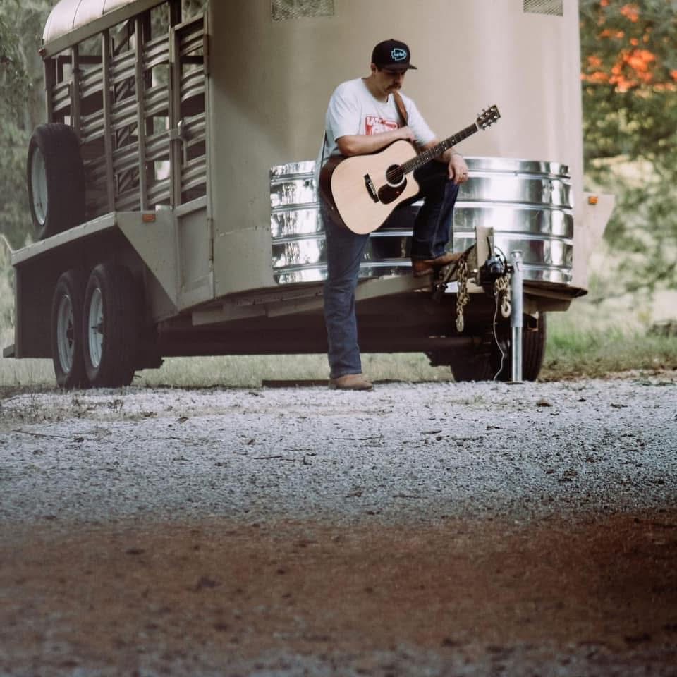 Person playing guitar outdoors near a trailer, standing on gravel with trees in the background.