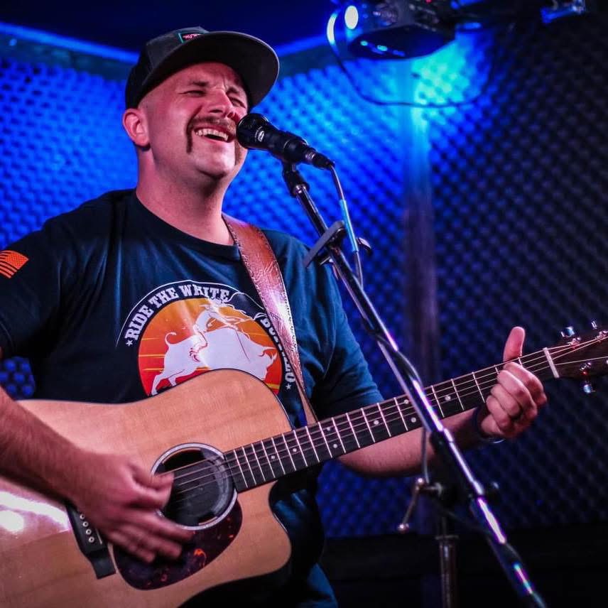 Person singing and playing guitar on stage with blue lighting and soundproof background.
