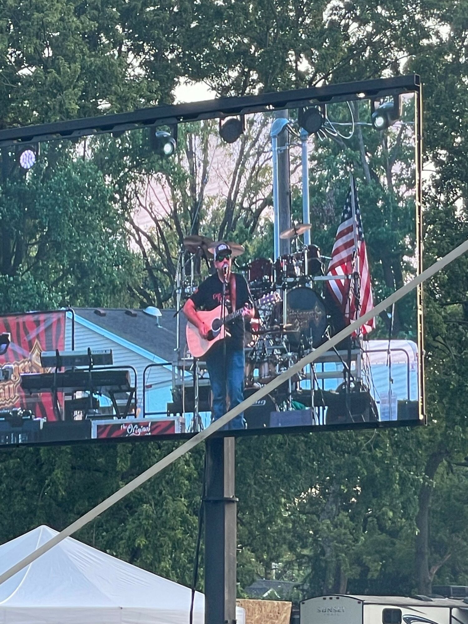 Musicians perform on an outdoor stage with trees in the background, American flag, and musical instruments, reflected in a large screen.