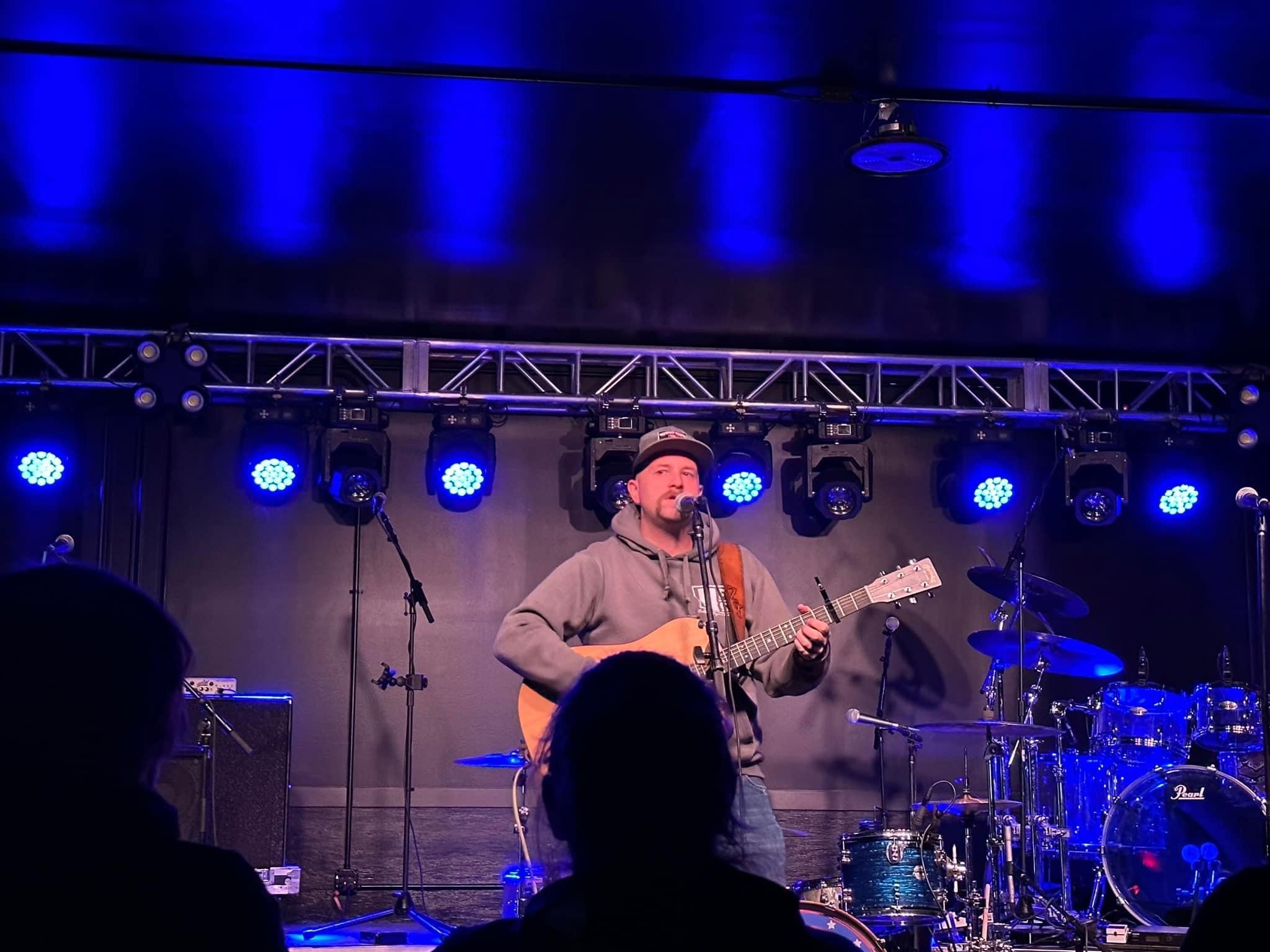 Person playing guitar and singing on stage with blue lighting and musical instruments, audience in foreground.