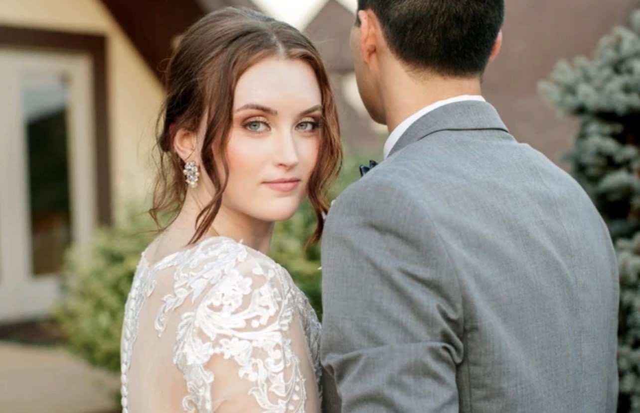 Woman with brown hair and earrings looks at camera, standing next to a man in a gray suit outdoors.