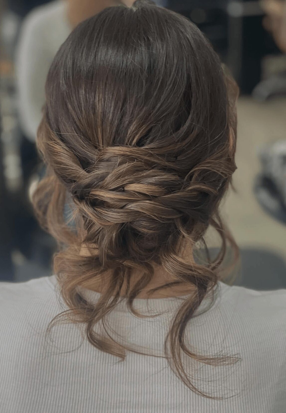 Back view of a woman with styled, wavy hair in an updo with loose curls, in a neutral setting.