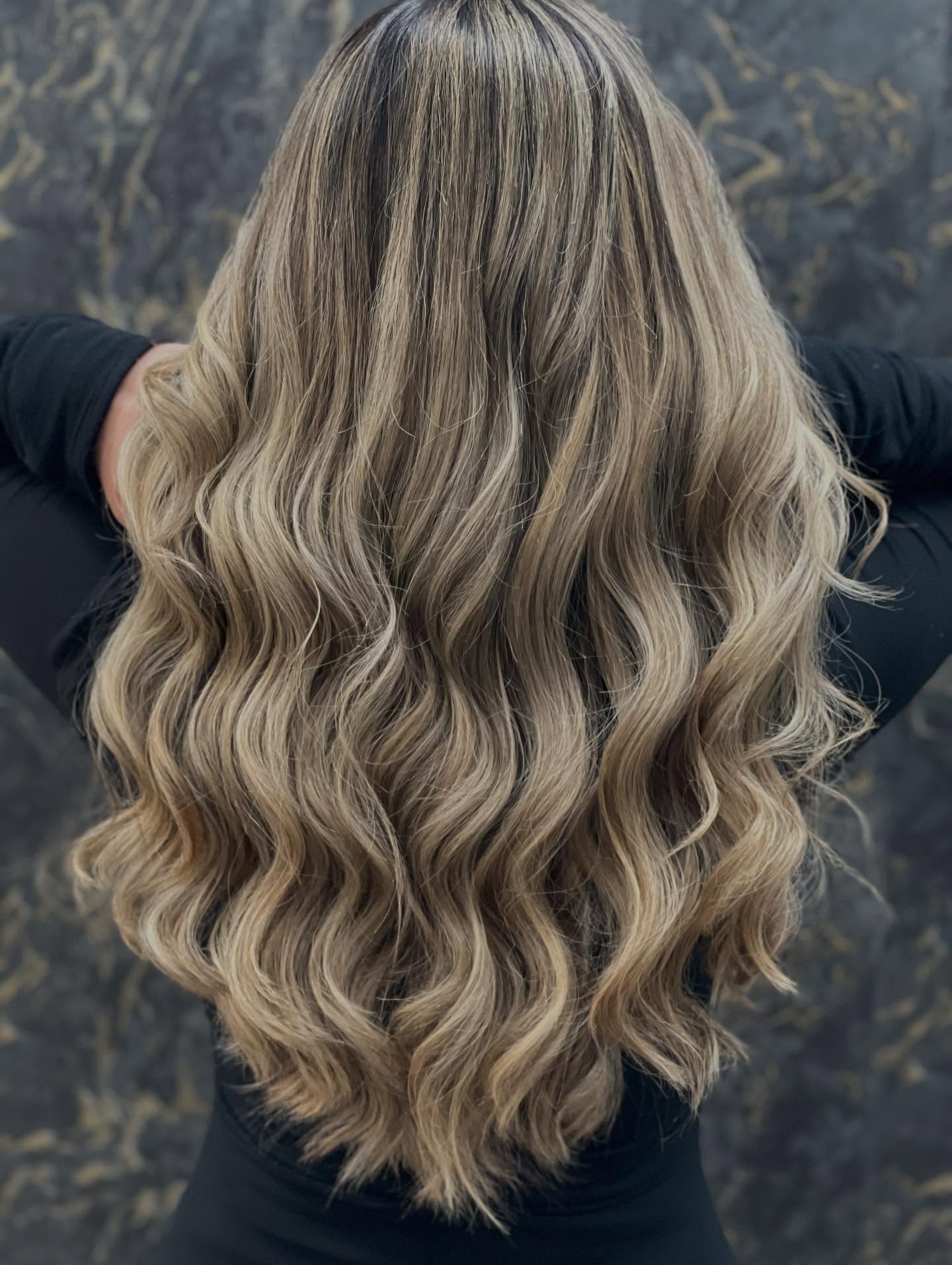 Back view of long, wavy blonde hair with darker roots, styled in loose curls, against a dark background.