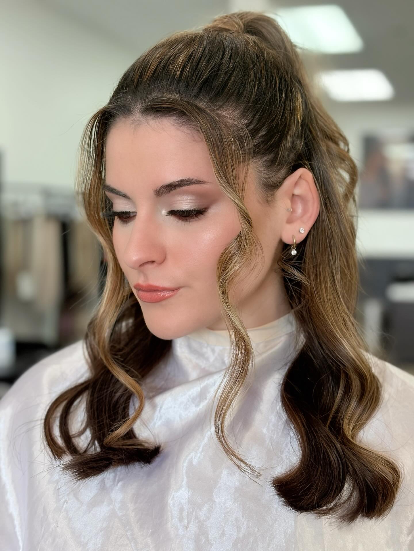 Young woman with long, wavy hair styled in a high ponytail, wearing a white top, in an indoor setting.