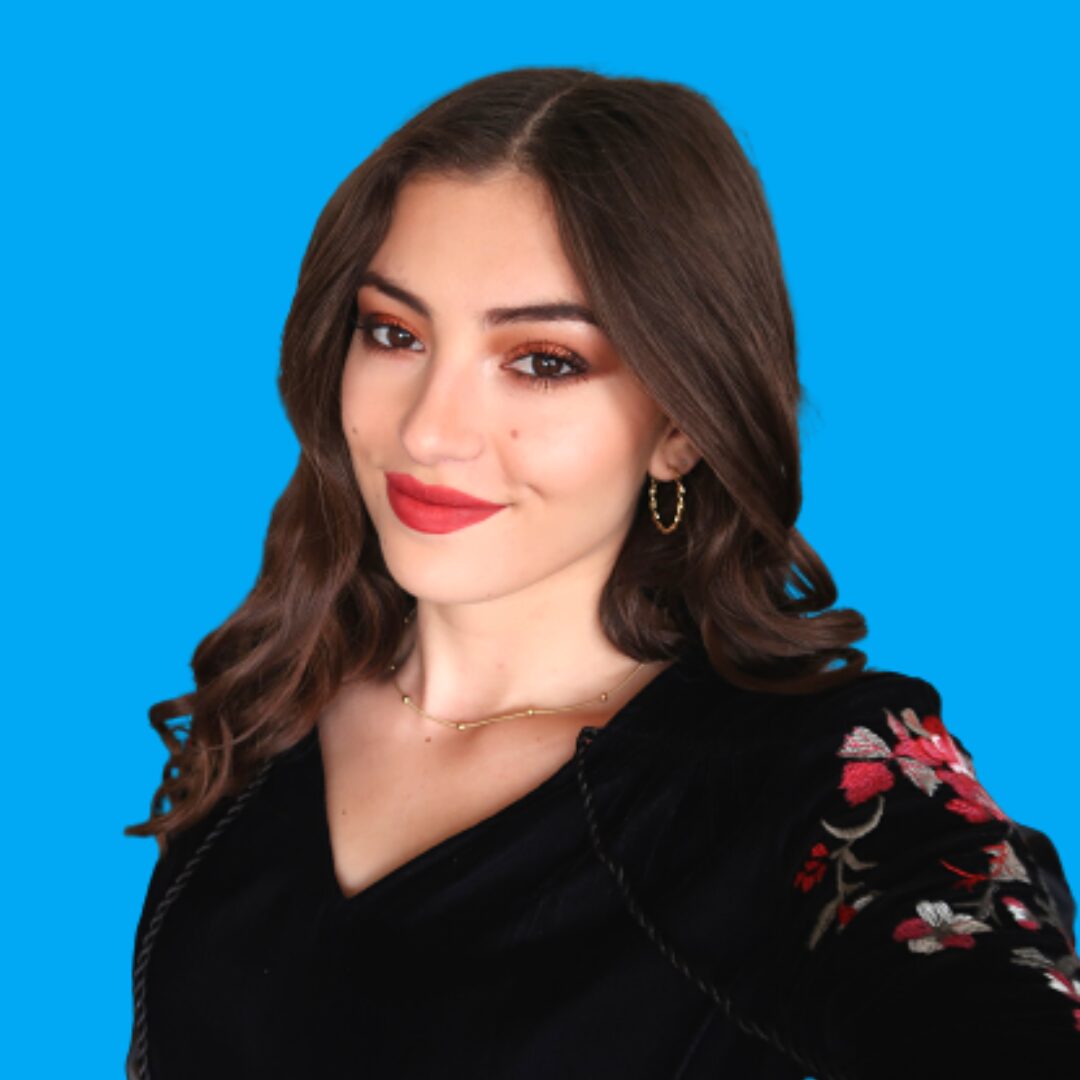 Young woman with long wavy brown hair, makeup, red lipstick, wearing a black top with floral embroidery, smiling, blue background.