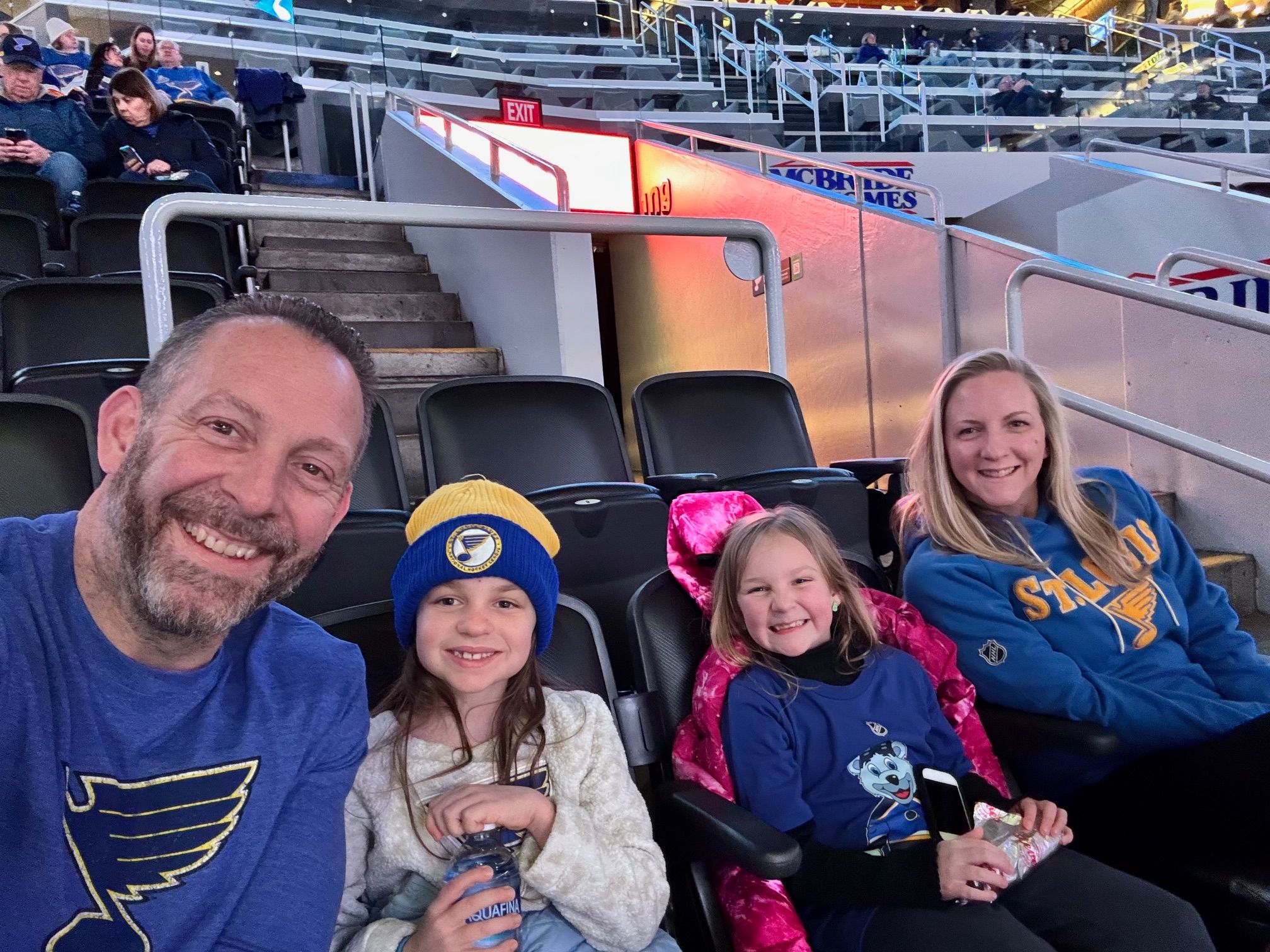 Four people sitting in stadium seats, smiling, at a sports event, with a stadium background.