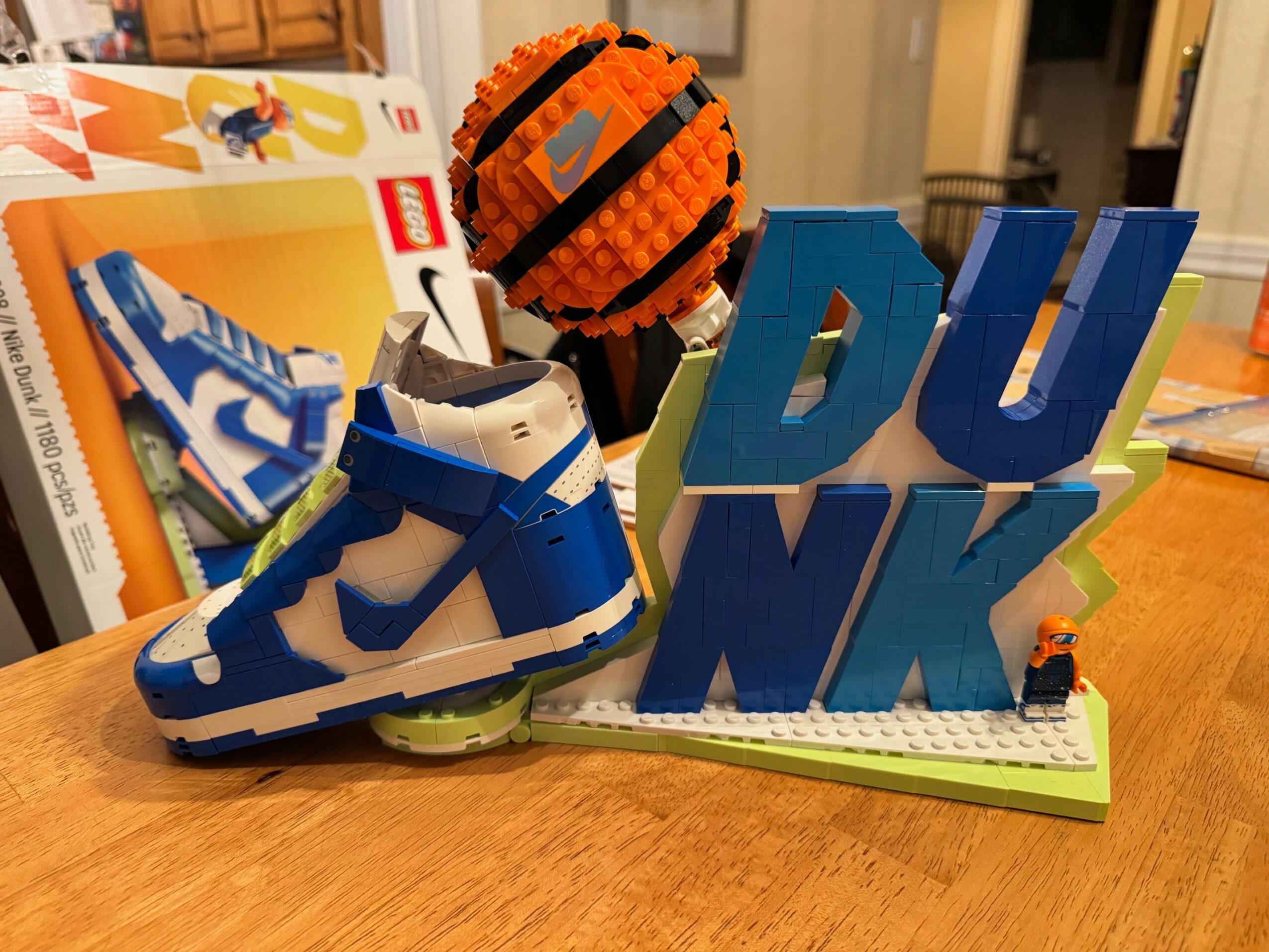 Sneaker, orange ball, and blue letters spelling DUNK on a table, with books and a room in the background.