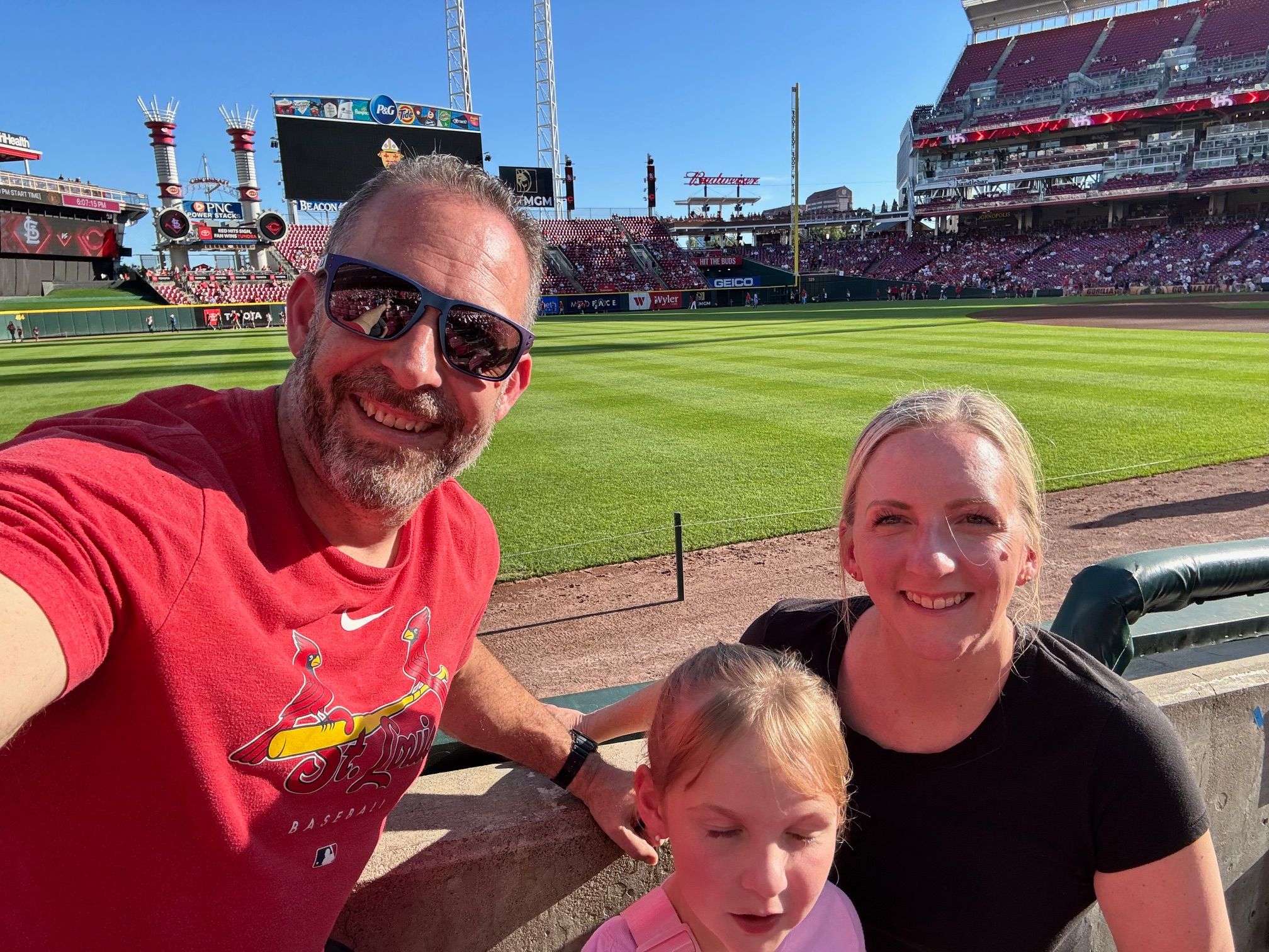 Three people smiling at a baseball stadium, with a field and stands in the background.