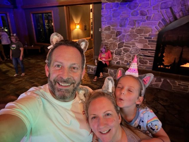 Three people wearing animal ears and party hats, smiling indoors with a stone fireplace in the background.