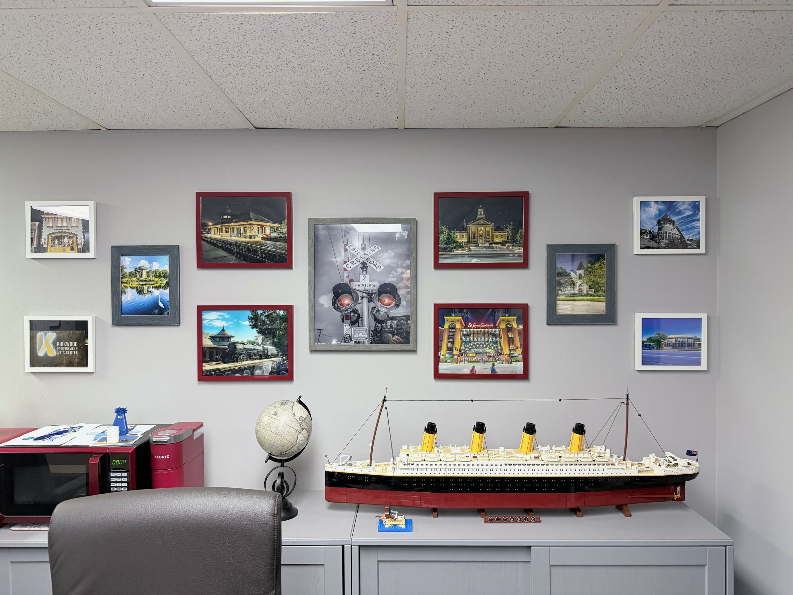 Wall with framed pictures and a model ship on a table, with a desk and chair in the foreground.