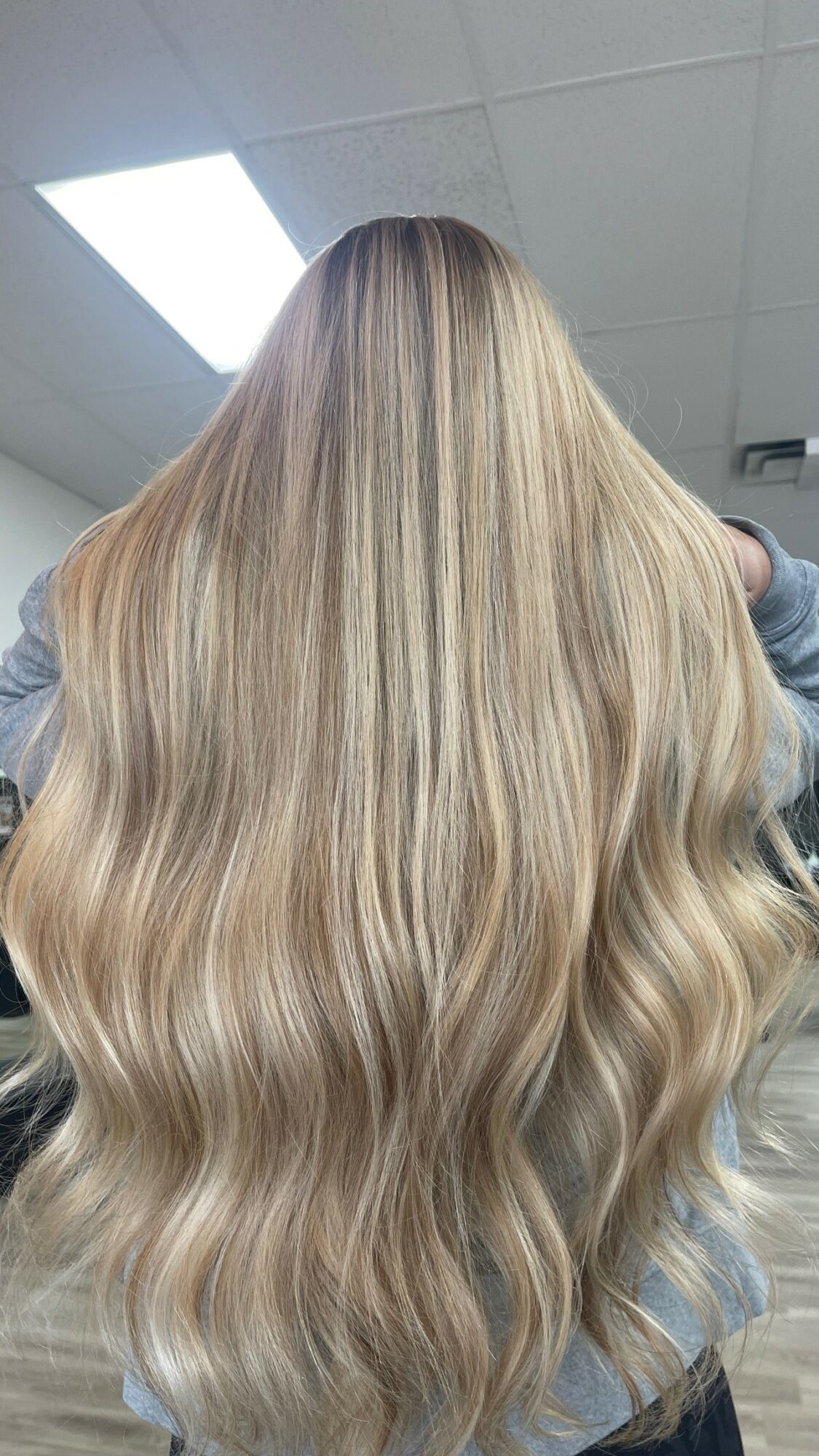 Back view of long, wavy blonde hair with highlights, indoors with ceiling lights visible