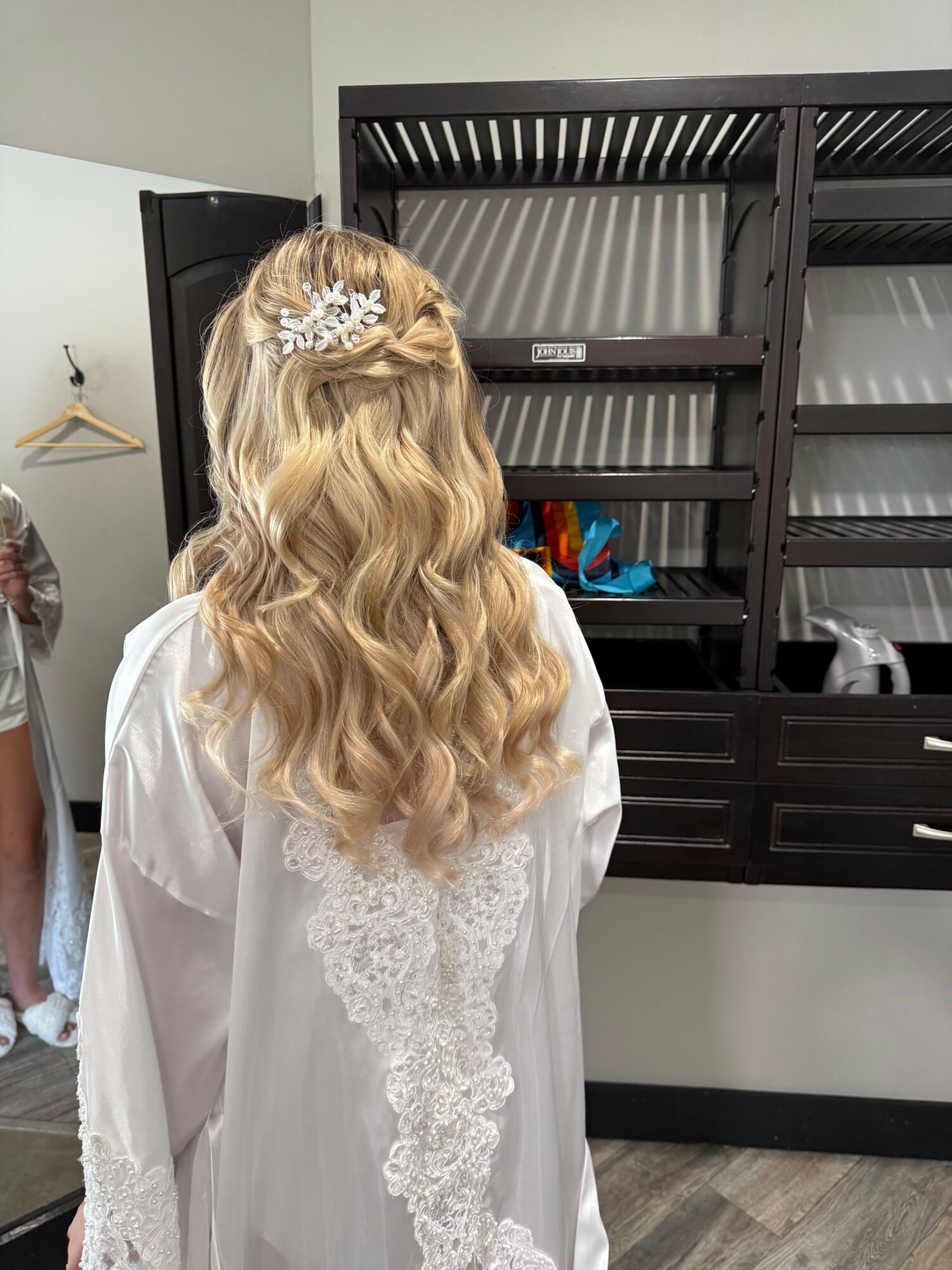 Back of a woman with long, wavy blonde hair wearing a white robe, standing indoors near a black shelf.