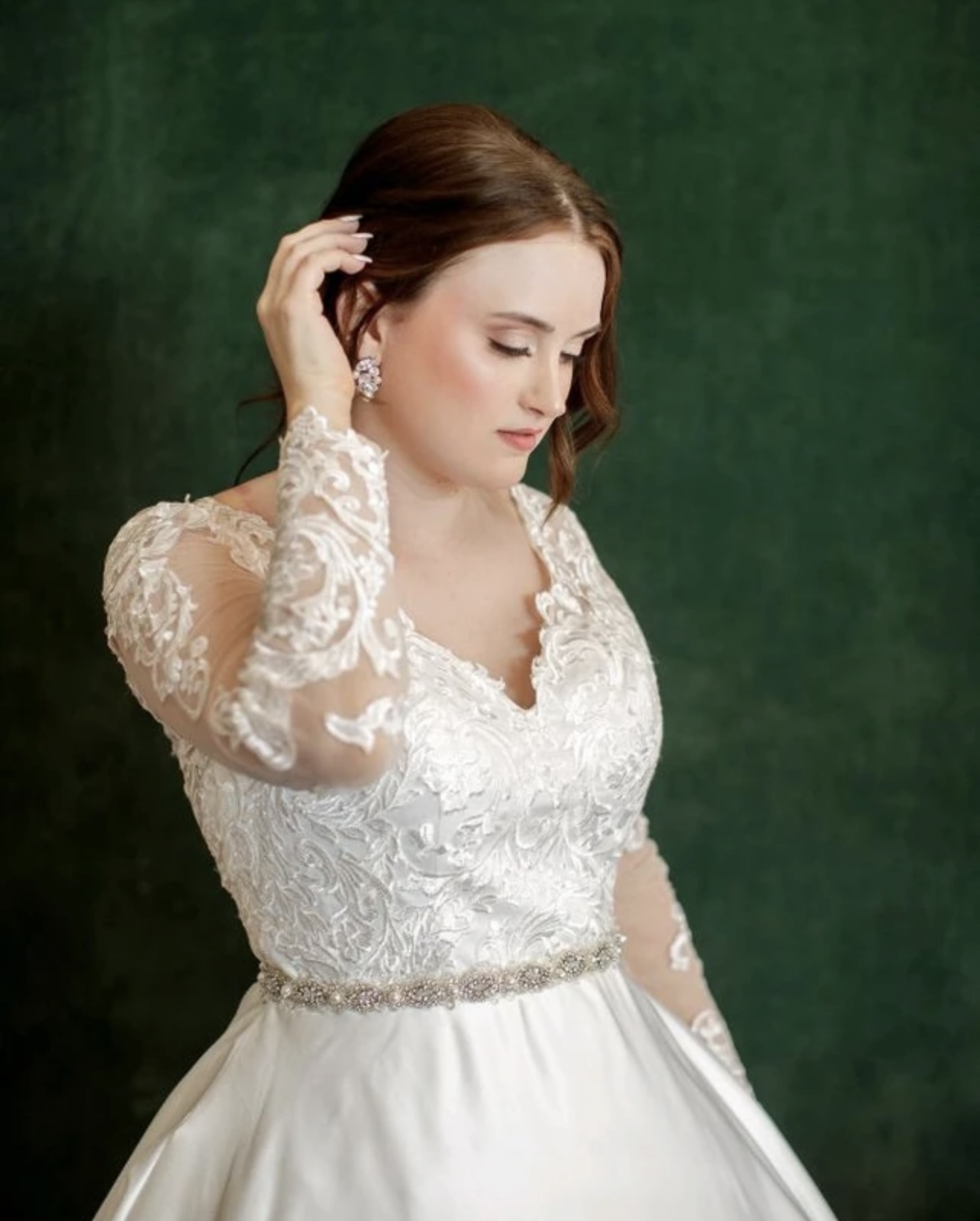 Woman in wedding dress with lace details, looking down, touching her hair, against a green background.
