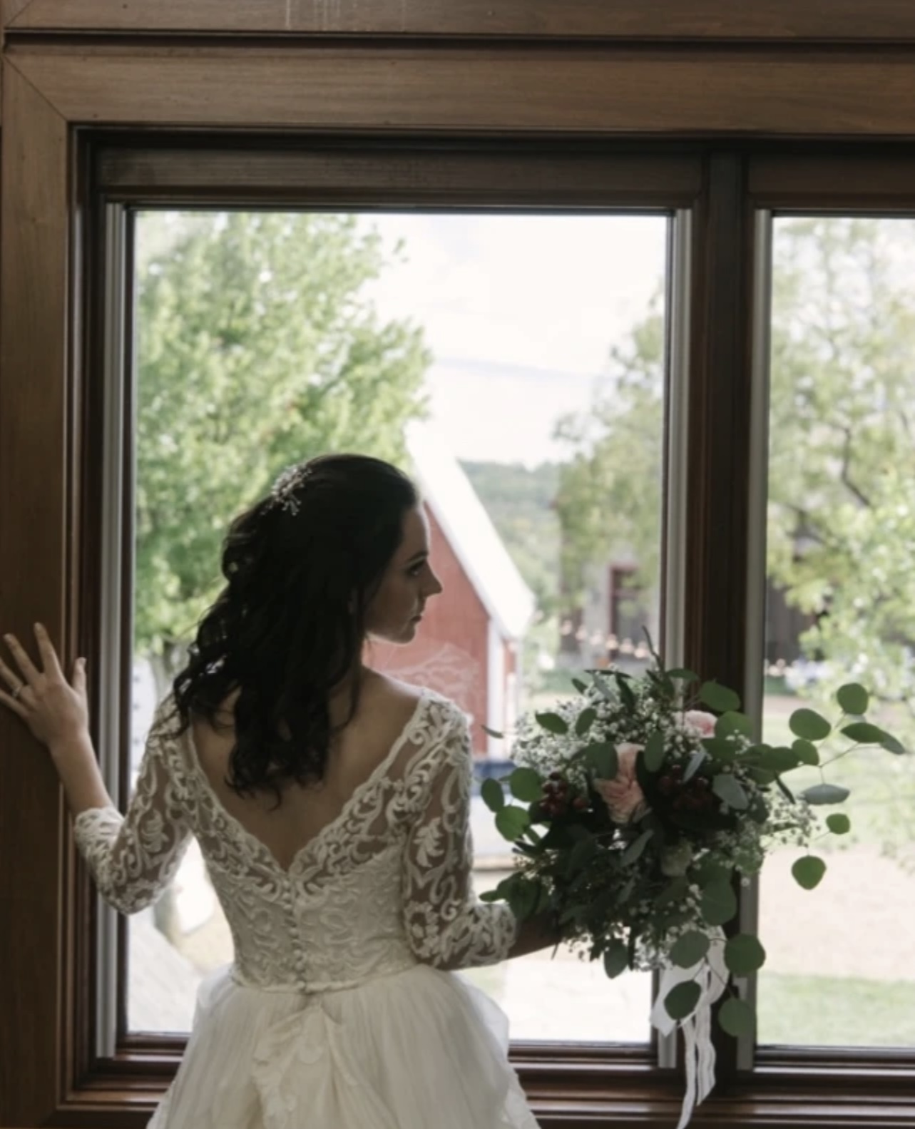 Woman in wedding dress holding bouquet, looking out window, outdoor scene with trees and buildings, natural light, lace dress, dark hair