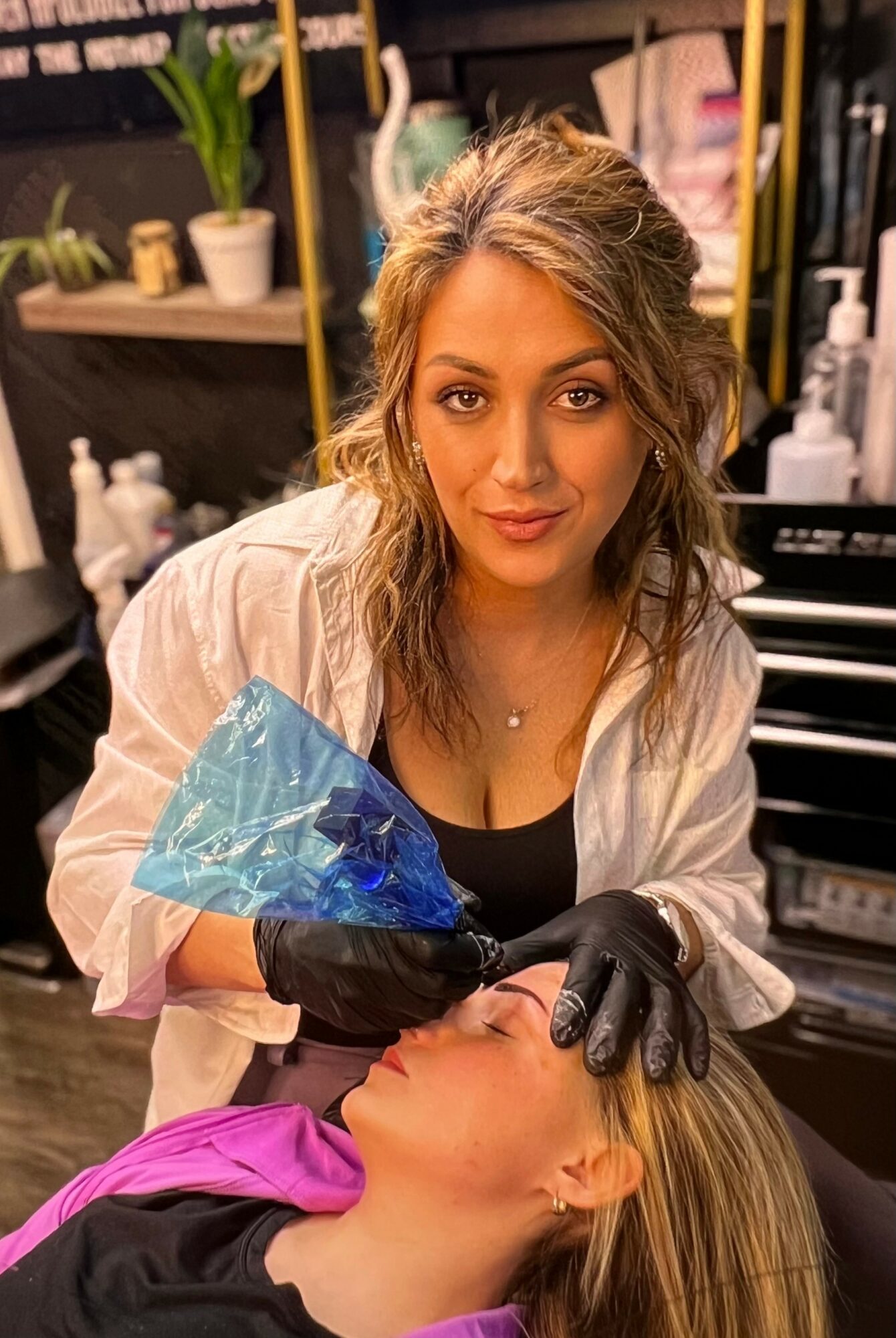 Woman with curly hair in a white coat holds a blue tattoo machine over a person's face in a tattoo studio.