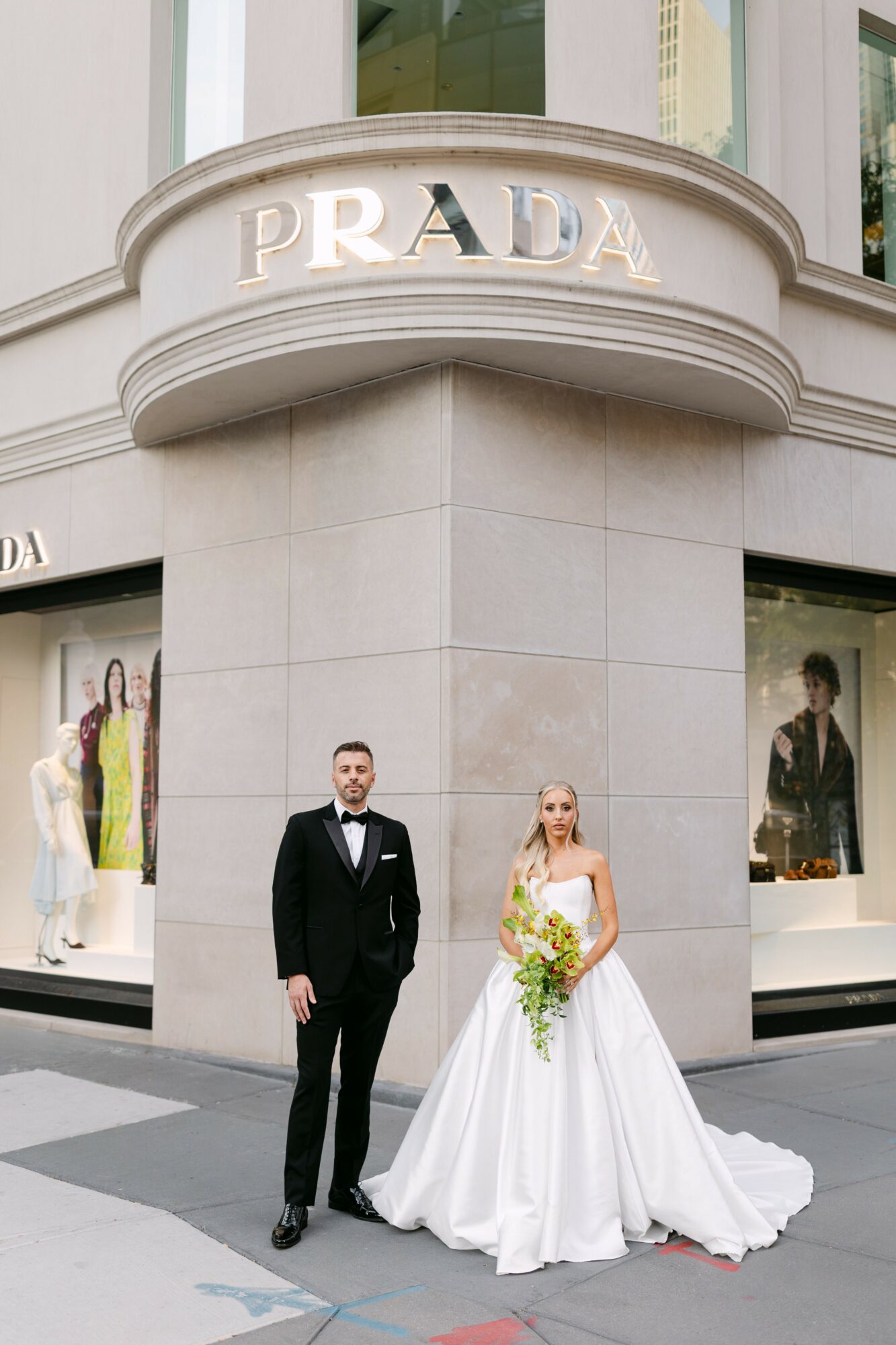 A man in a tuxedo and a woman in a wedding dress stand outside Prada store, holding a bouquet of flowers.