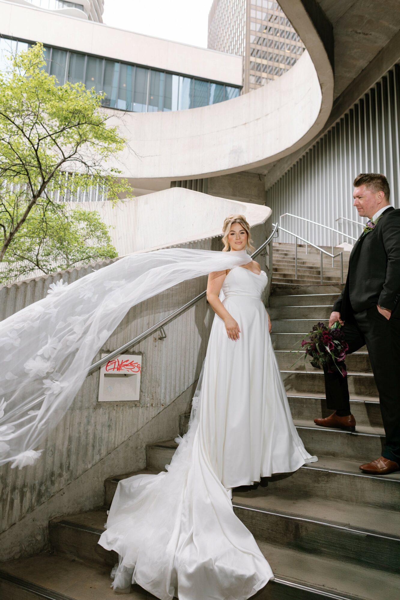 Bride in white dress and long veil stands on stairs, groom in black suit holds flowers, modern building background.