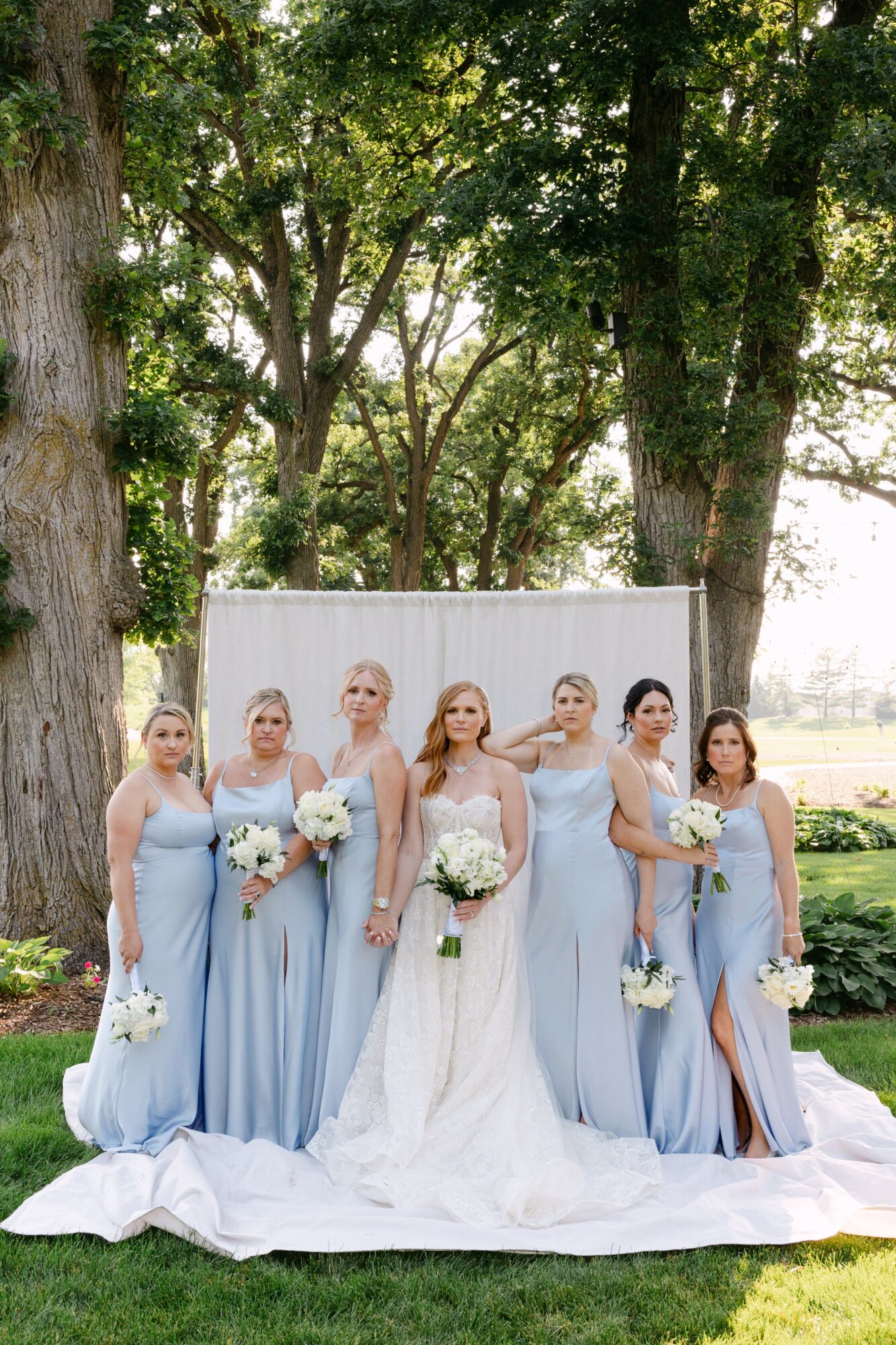 Seven women in light blue dresses holding white bouquets, standing outdoors in front of trees and a white backdrop.
