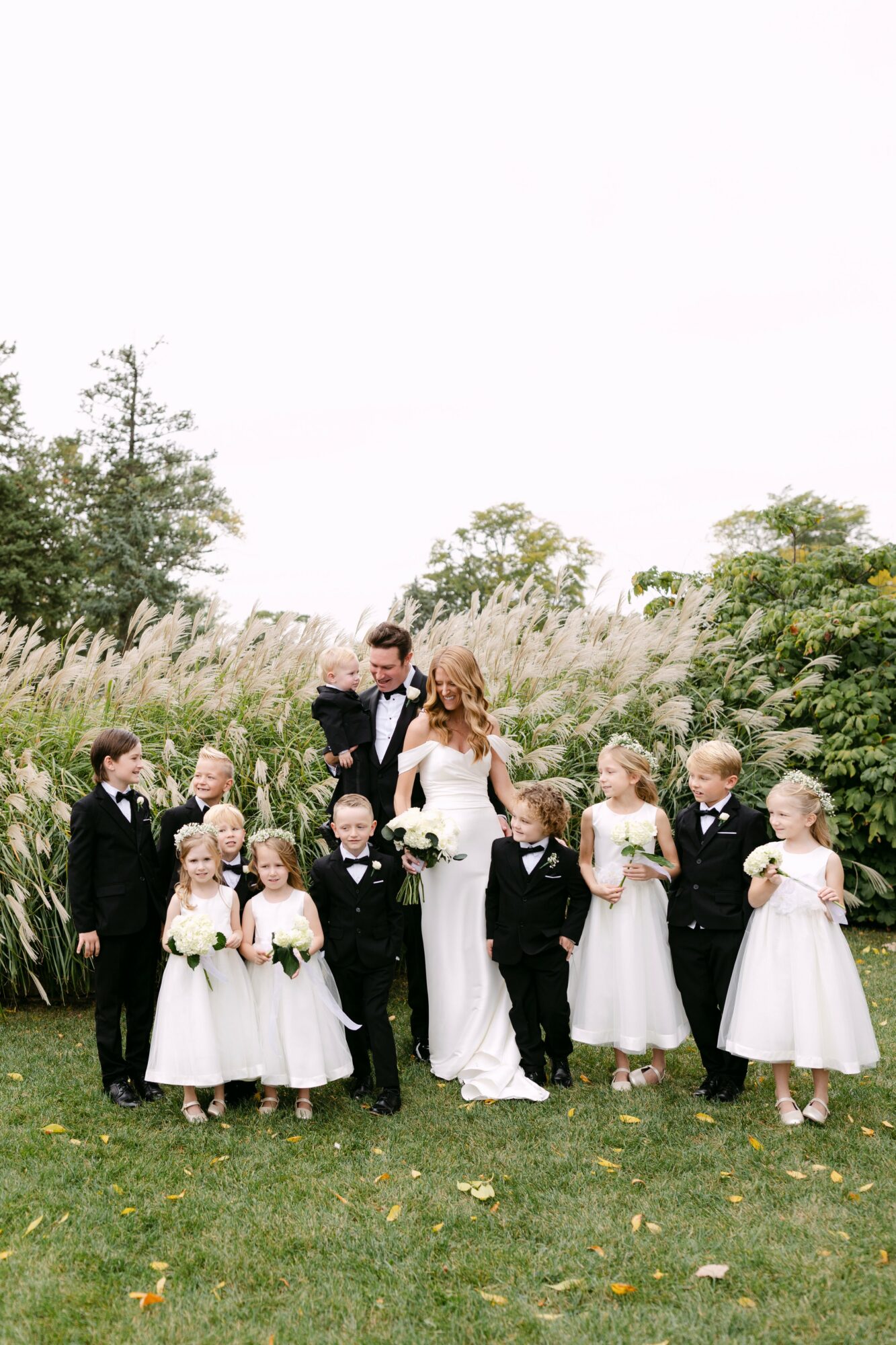 A bride in a white dress and a groom in a tuxedo stand outdoors with children in formal attire, greenery in background.